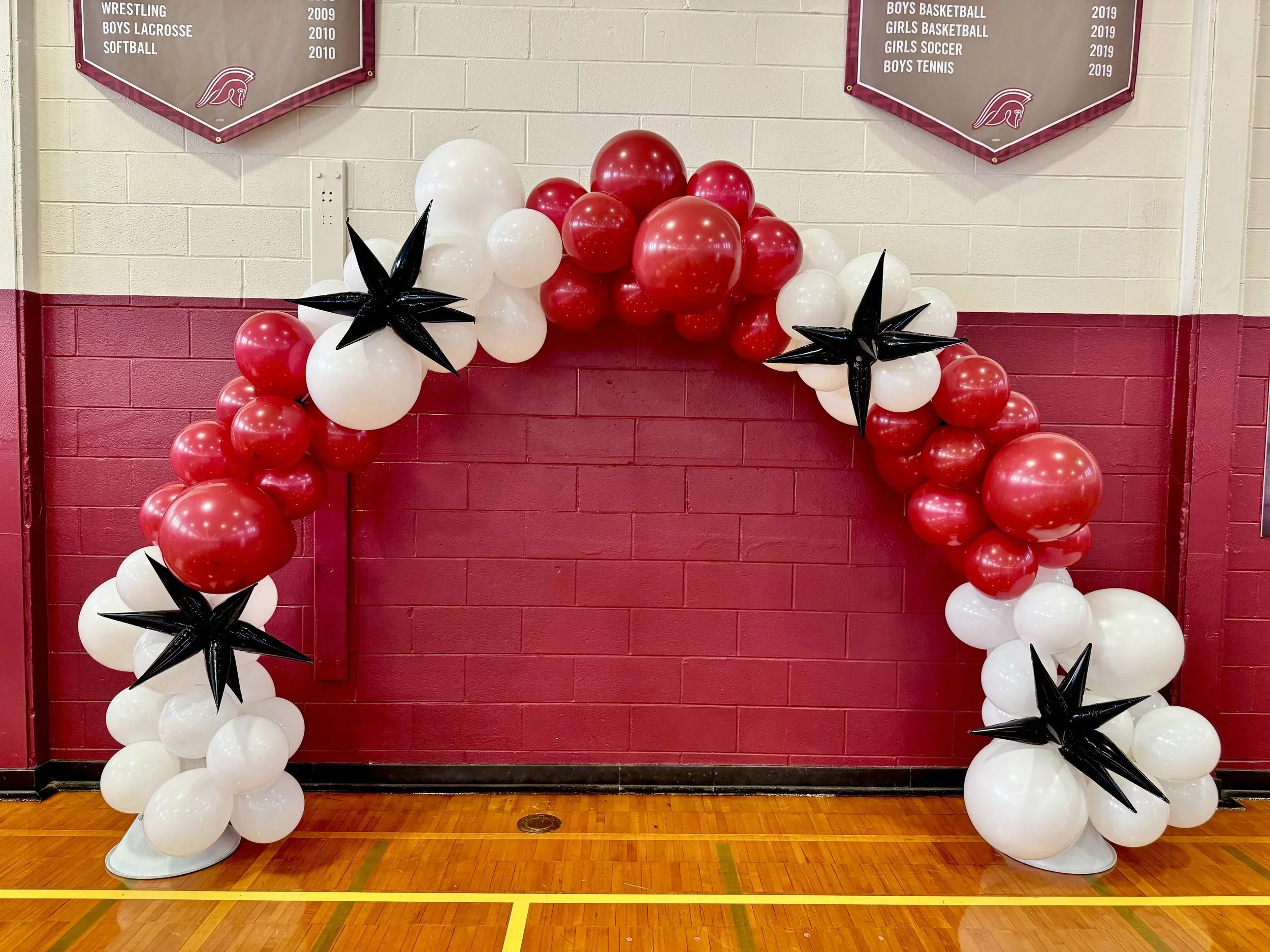 Decorative basketball arch made of white, red, and black balloons in a gymnasium, with banner plaques listing achievements.