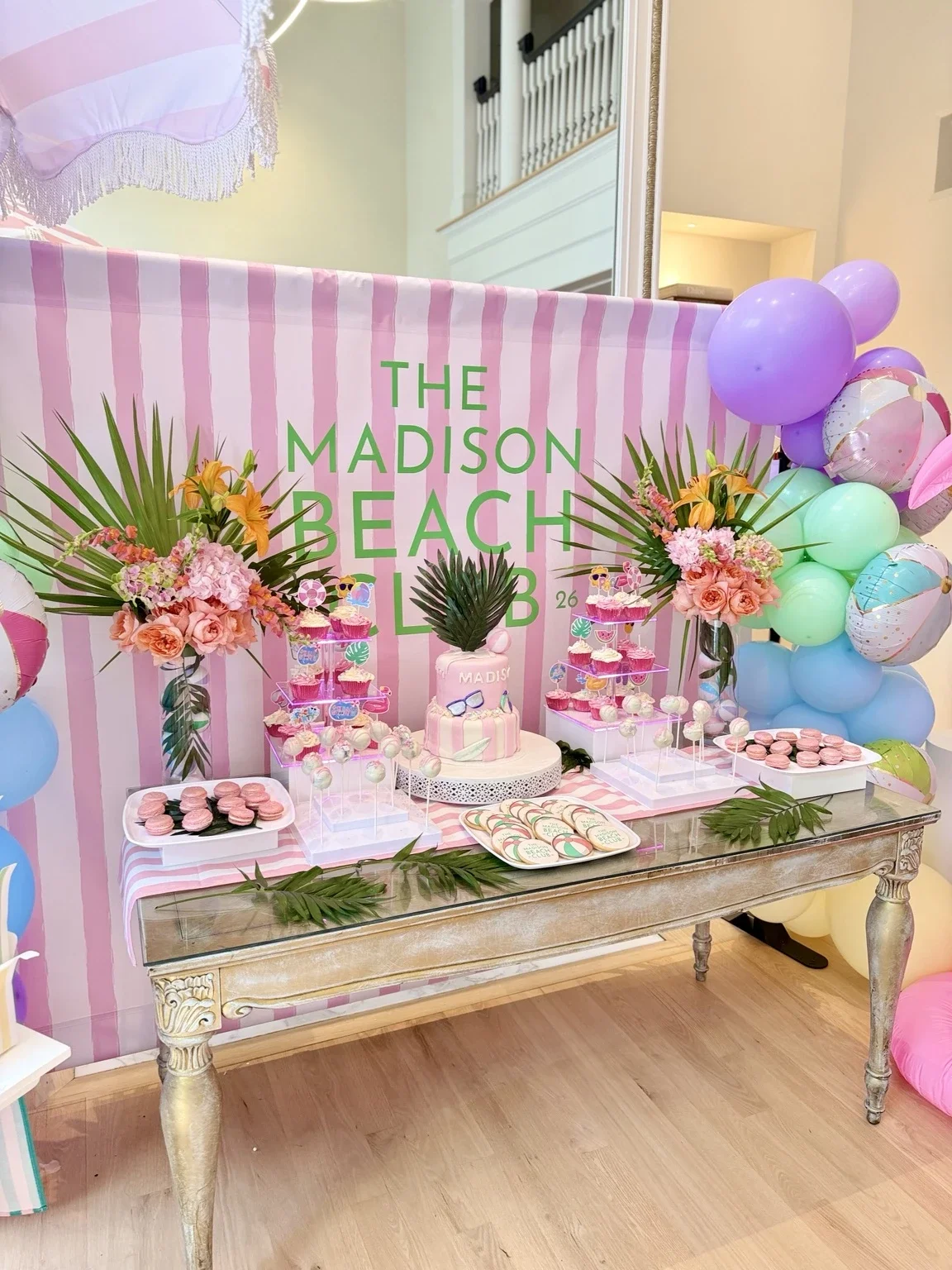 Luxury beach club themed 6th birthday dessert table with custom cake, pastel balloon garland, tropical floral arrangements, cupcakes, macarons, and pink striped backdrop designed by Stellar View Events in New Jersey.