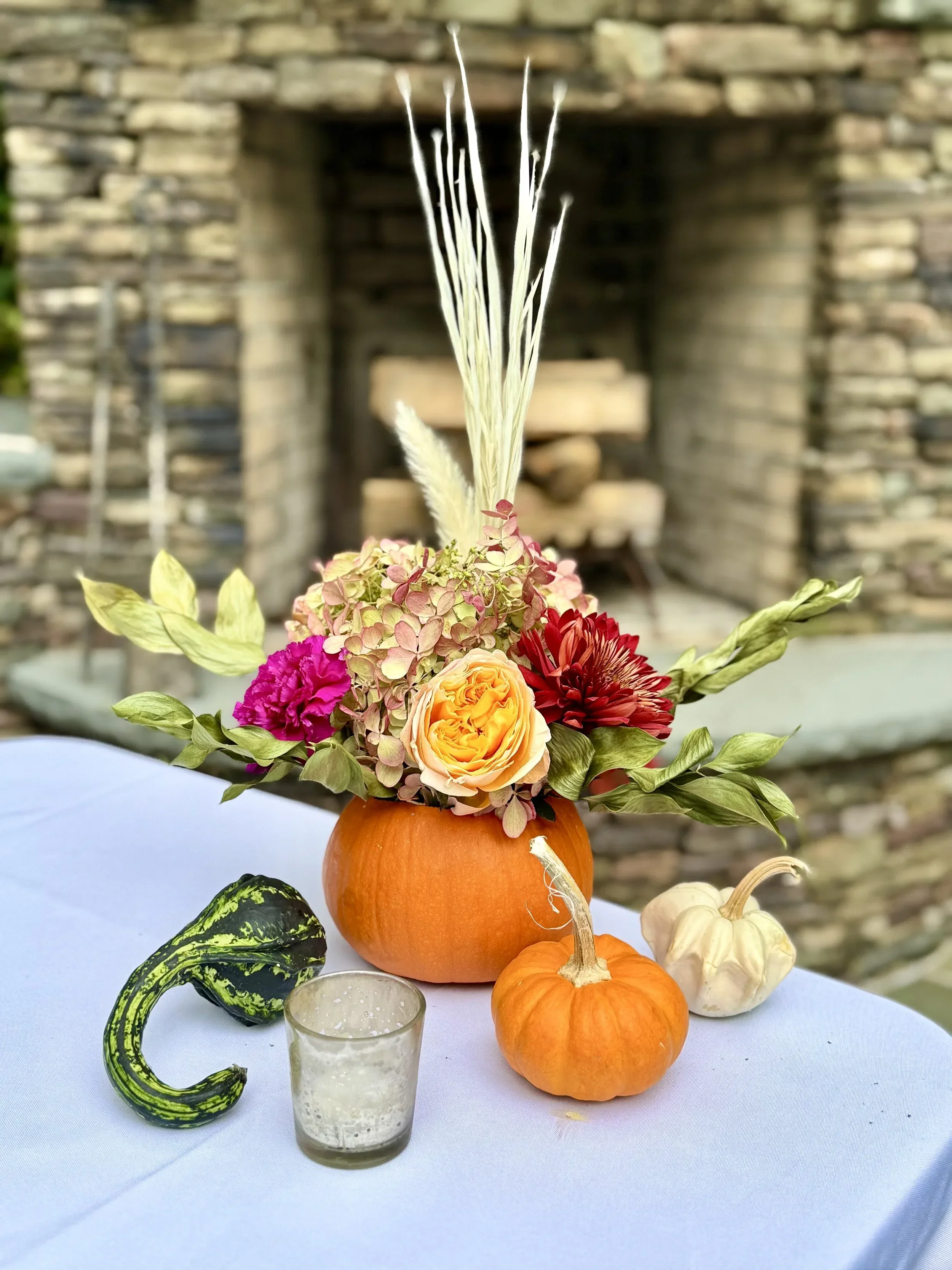 A pumpkin filled with pink, yellow, red, and purple flowers, with decorative wheat stalks sticking out from the middle, placed on a white tablecloth with smaller pumpkins, gourds, and a glass votive candle around it, set outdoors with a stone firepla