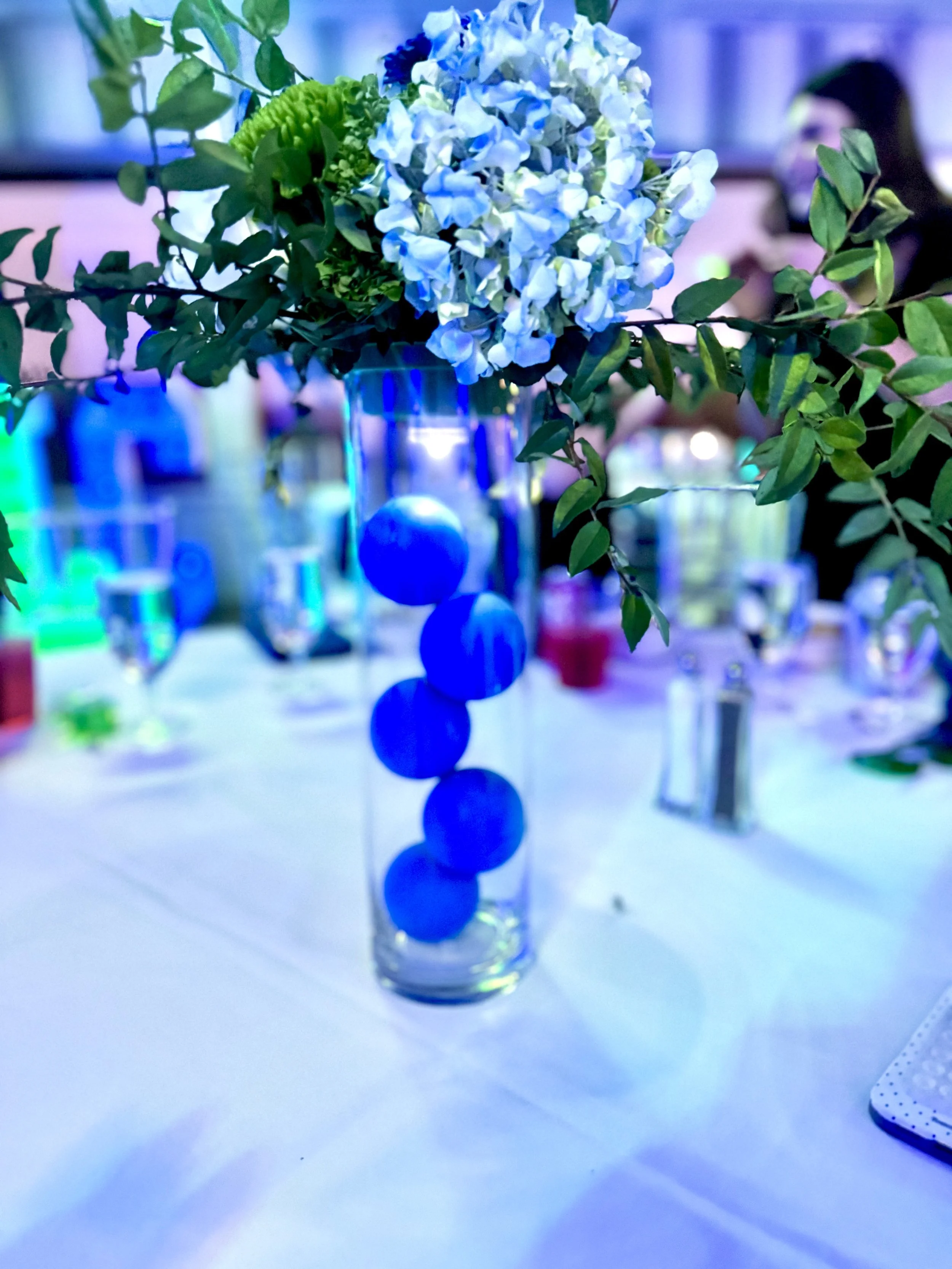 Decorative floral centerpiece featuring white and blue flowers with green leaves in a tall glass vase, arranged on a table at a formal event.
