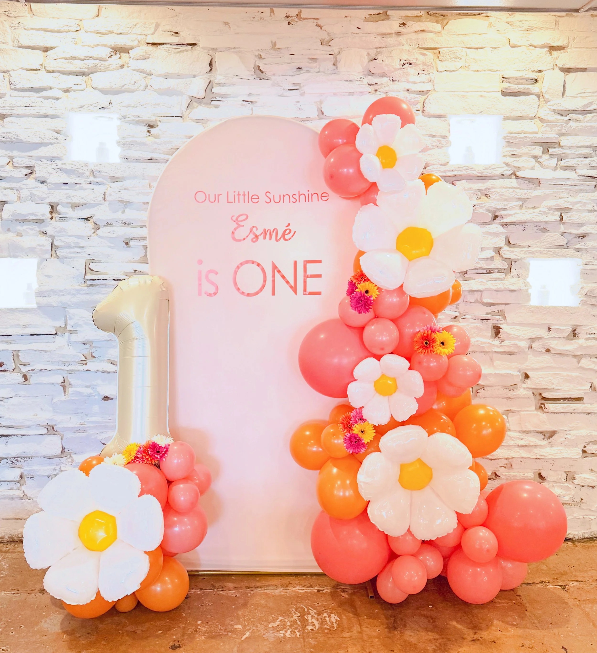 Decorative arrangement of pink, white, and orange balloons with artificial flowers, set against a white brick wall, with a backdrop that reads 'Our Little Sunshine Esmé is ONE'.