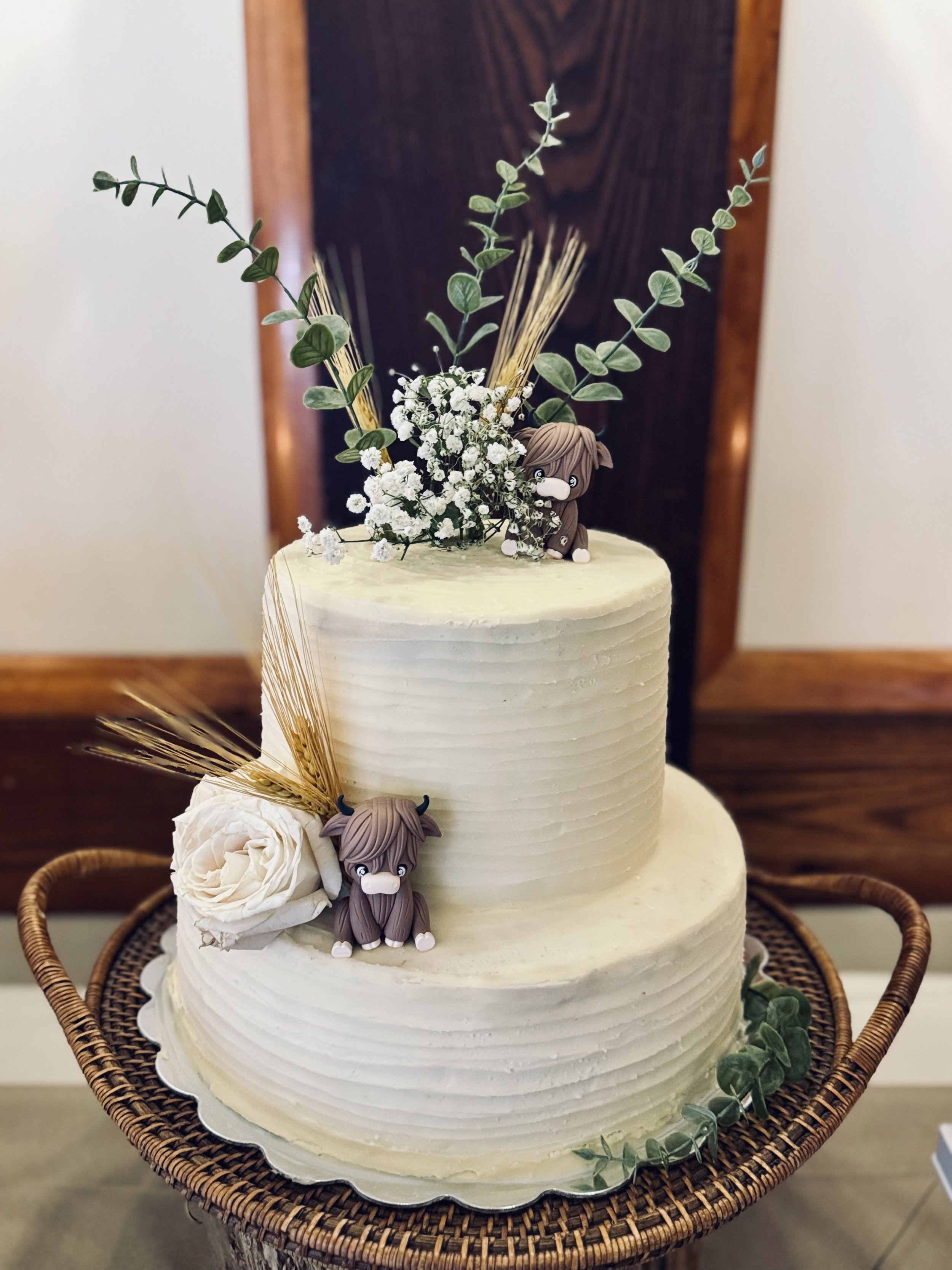 Two-tiered wedding cake decorated with greenery, small white flowers, a white rose, and two small animal figurines with horns and white noses, all placed on a wicker table. The cake has a smooth, textured white frosting.