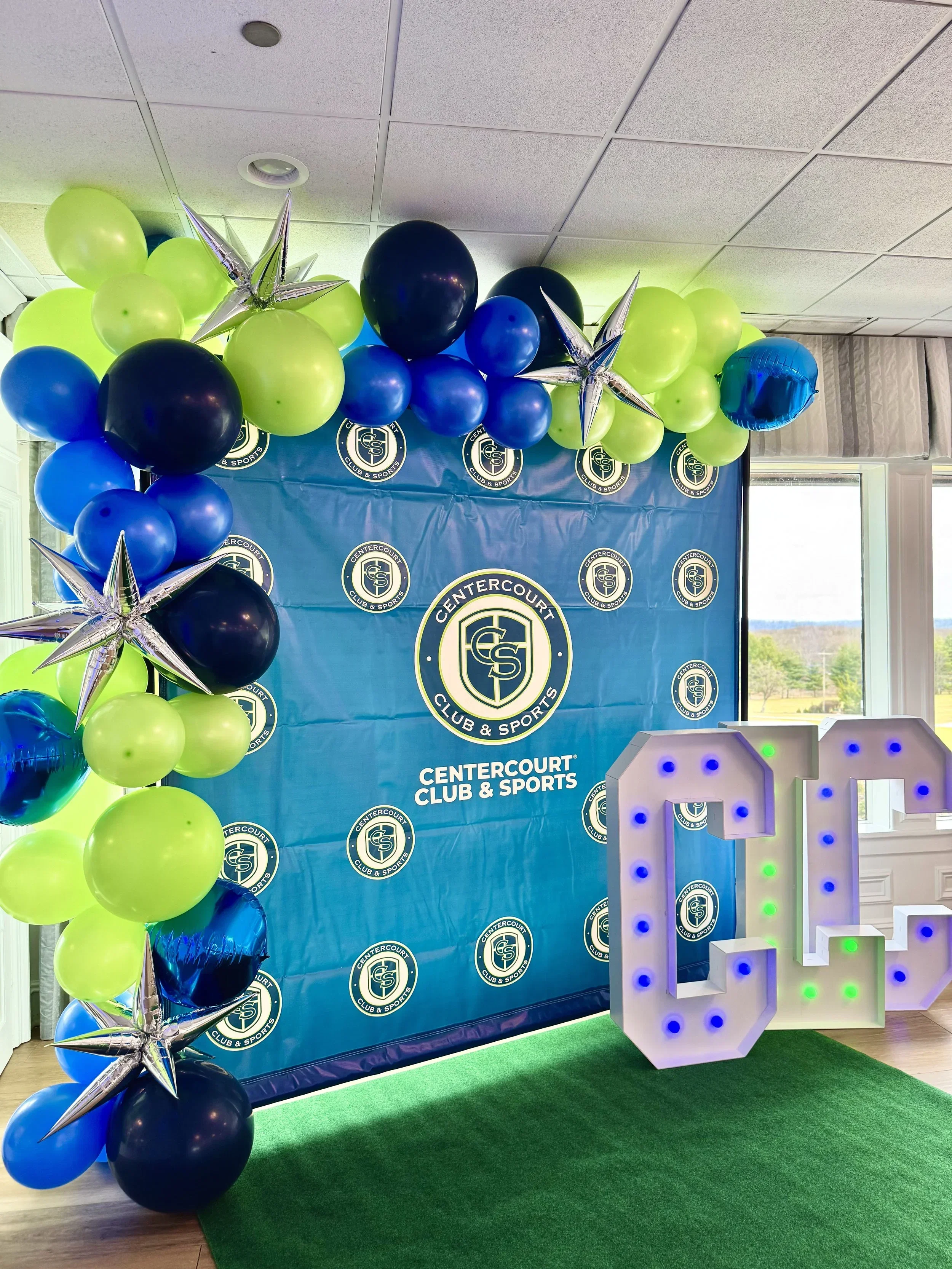 Decorative balloon arch in blue, green, and black with metallic star-shaped balloons, set against a blue backdrop with the CenterCourt Club & Sports logo, and a large illuminated letter D on a green carpet.