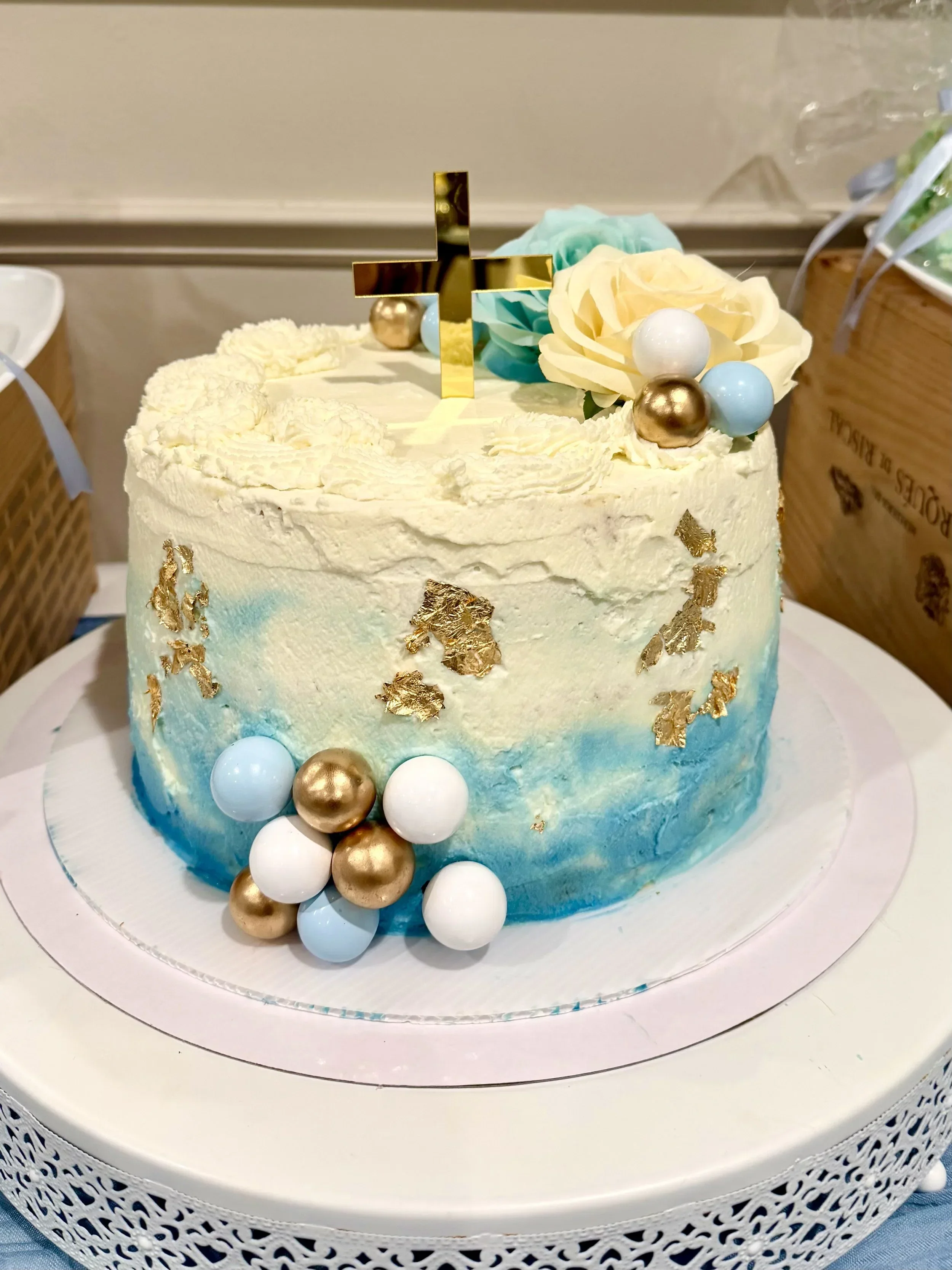 A decorated birthday cake with blue, white, and gold accents, topped with a metallic cross and floral decorations, sitting on a white cake stand.