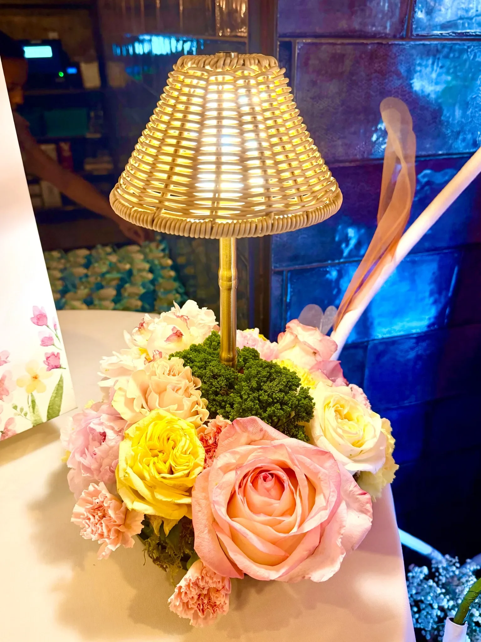 A floral arrangement with pink, yellow, and cream roses, pink carnations, green foliage, and a small table lamp with a wicker shade on a white surface, with a background of a dark wall and blue lighting.