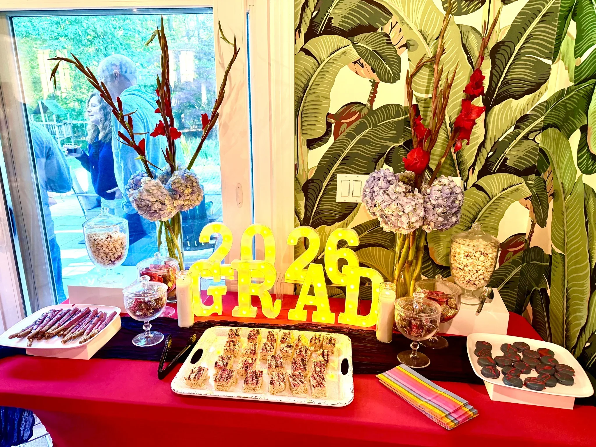 Table with various snacks such as popcorn, pretzels, brownies, and cookies. Decorative floral arrangements with large green leaves and red flowers. A bright yellow '2016' light-up sign is in the center. The background features a window and a wall wit