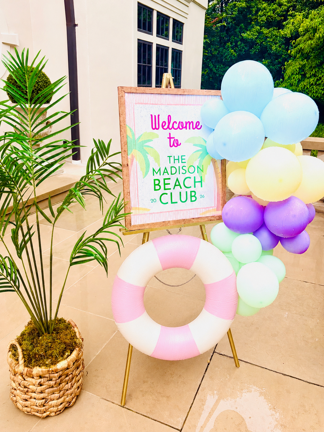 A welcome sign for the Madison Beach Club with pastel-colored balloons, a decorative life preserver, and a potted plant nearby on a patio.