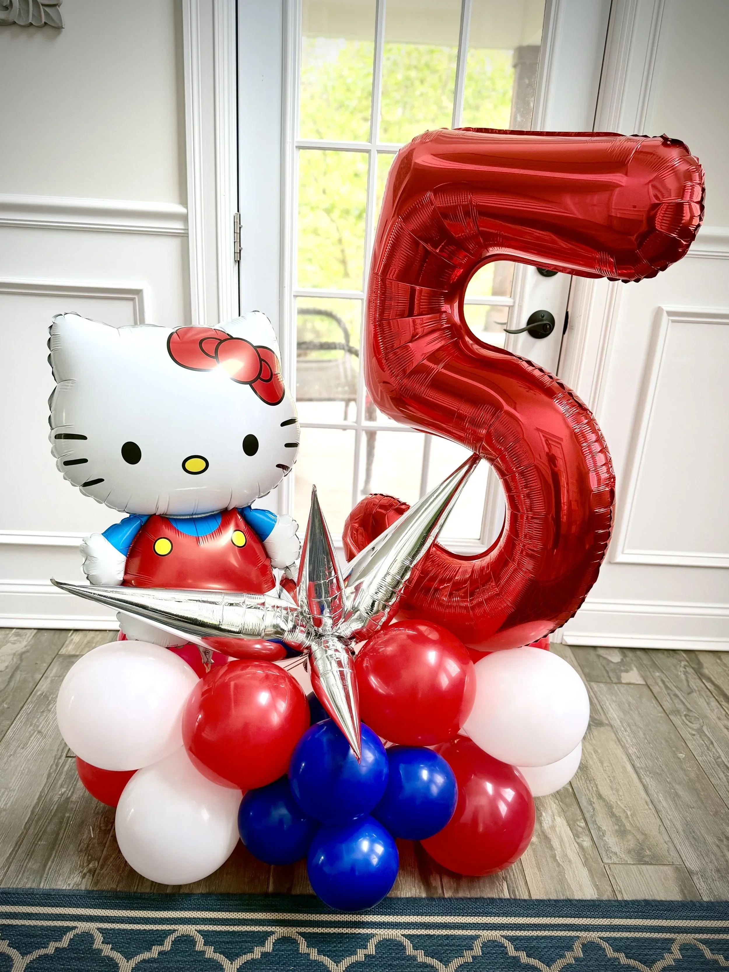 A colorful balloon arrangement featuring a large red number five balloon, a Hello Kitty balloon, and a cluster of red, white, and blue balloons, set indoors near a door with glass panels and a view of greenery outside.