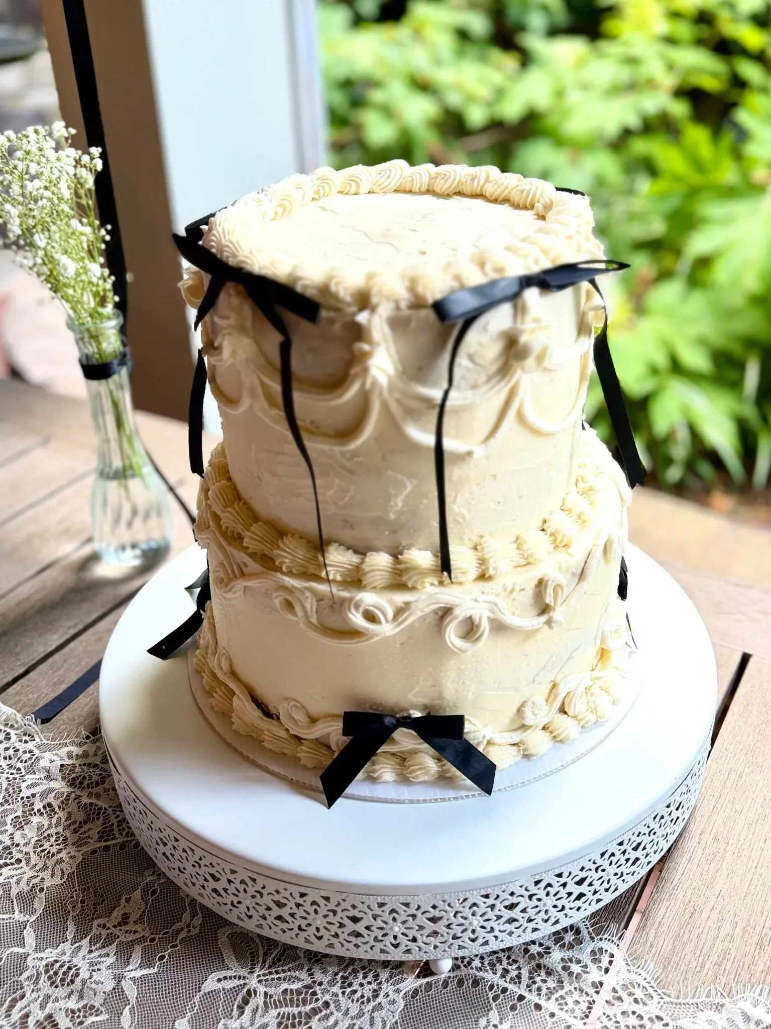 A white three-tiered cake decorated with white frosting, black ribbons, and ivory-colored piping, placed on a white decorative cake stand on a wooden table with a lace tablecloth. There are green plants visible outside a window in the background.