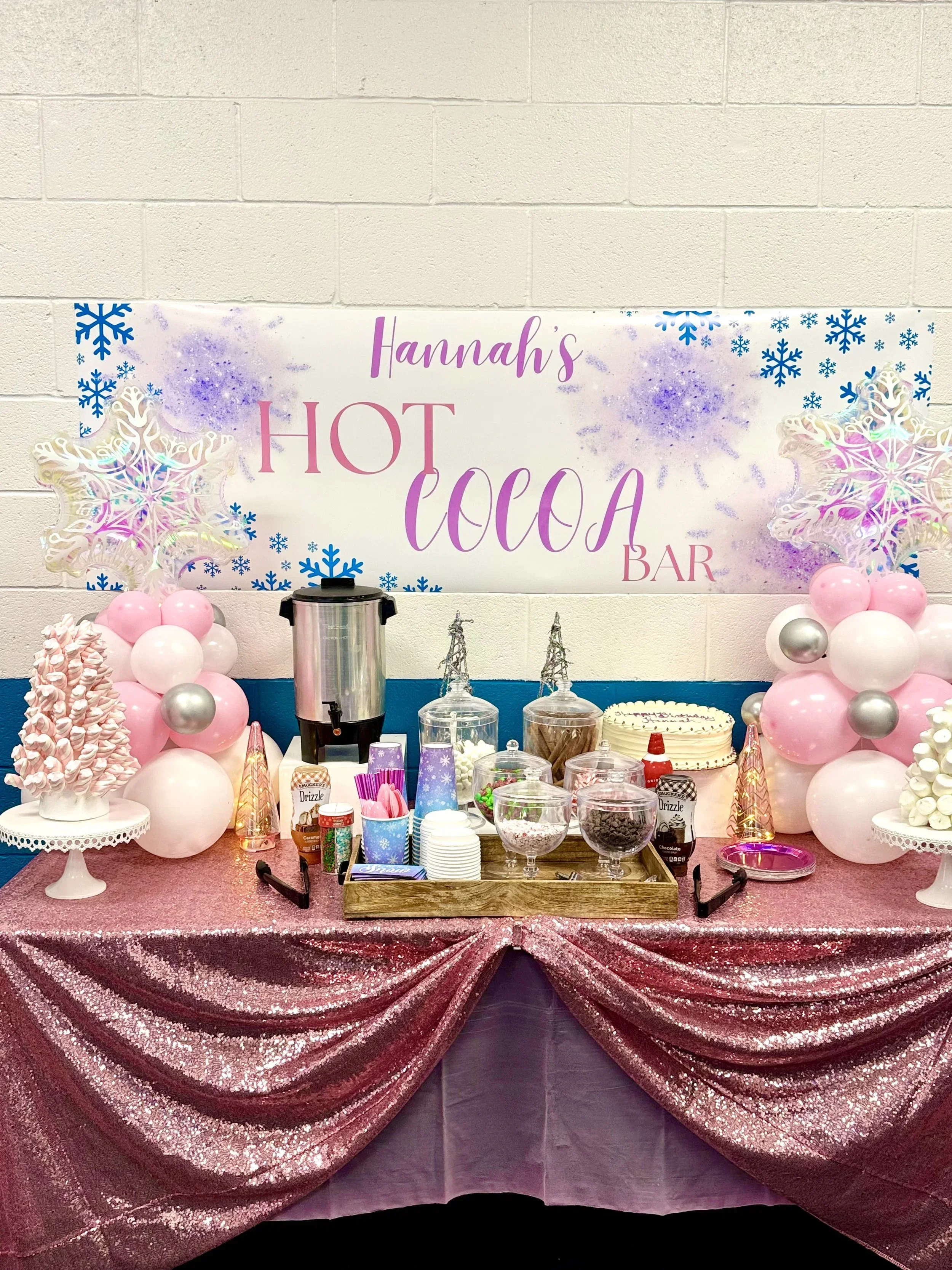 Decorated party table with pink and silver balloons, large snowflake decorations, pink glittery tablecloth, various candies, popcorn, and drinks, with a sign reading 'Hannah's Hot Cocoa Bar' in the background.