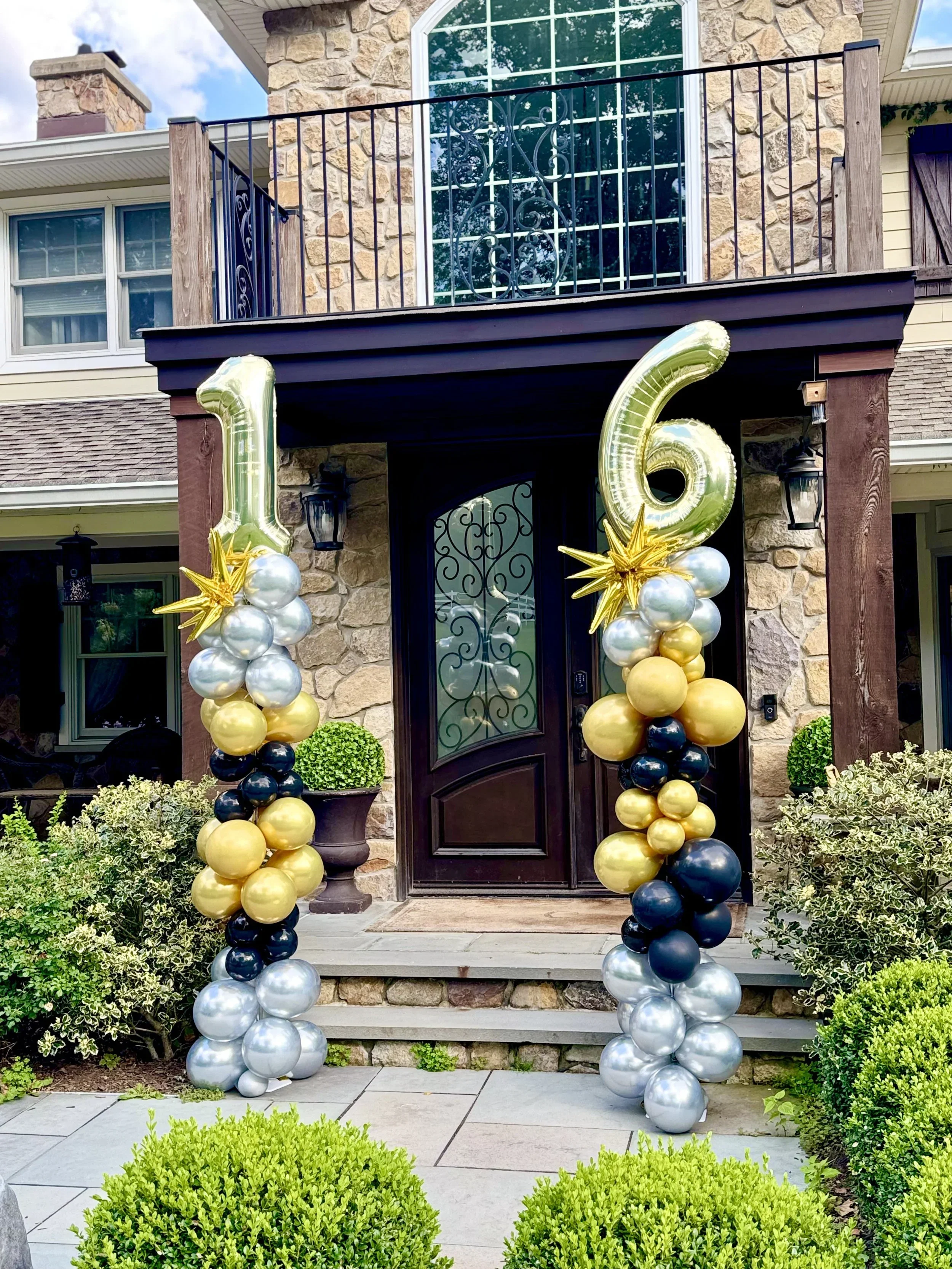 Front porch decorated with silver, gold, and black balloons forming the number 16, signifying a celebration, likely a 16th birthday or anniversary, with greenery and a stone house in the background.