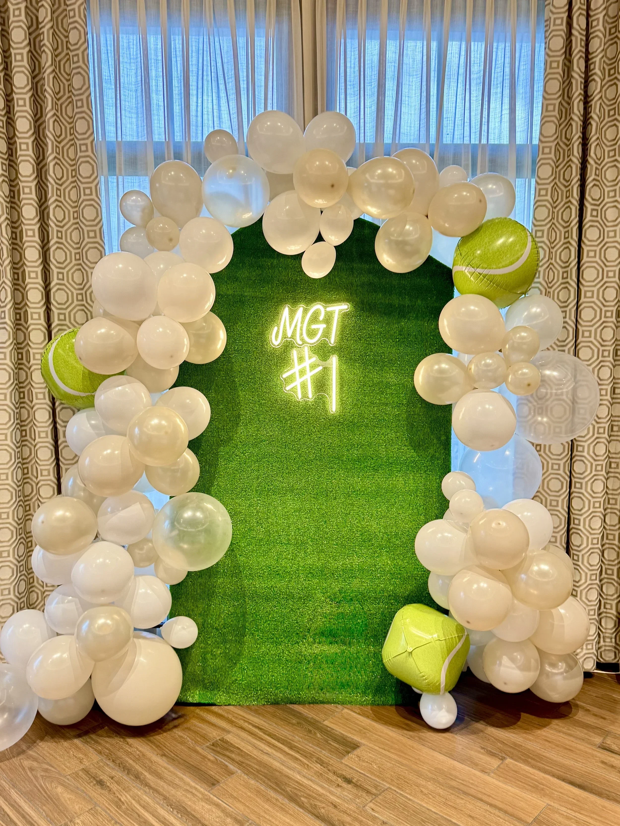 Decorative balloon display with white balloons of various sizes and a large green tennis-themed balloon, set against a green grass-like backdrop with a neon sign reading 'MGT #1'.