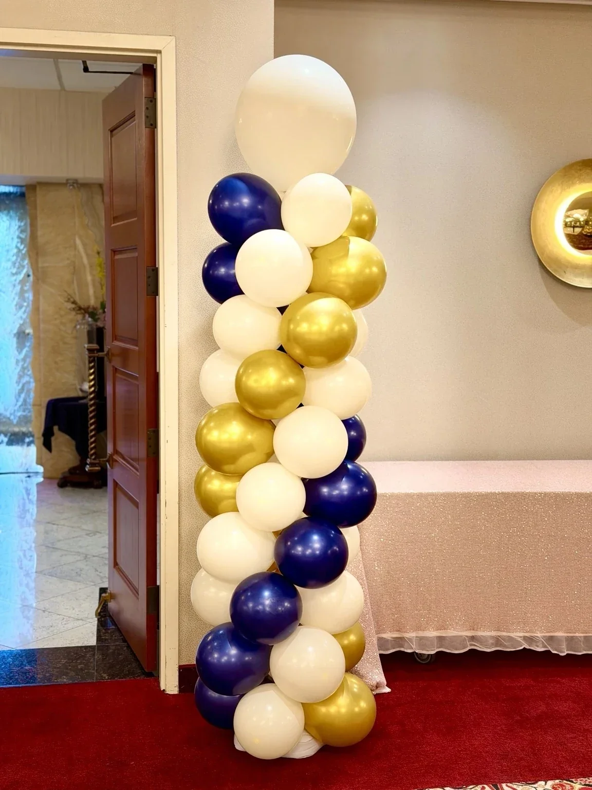 A tall column of balloons in colors white, navy blue, and gold, arranged in a vertical pattern, next to a wall and doorway in an indoor setting.
