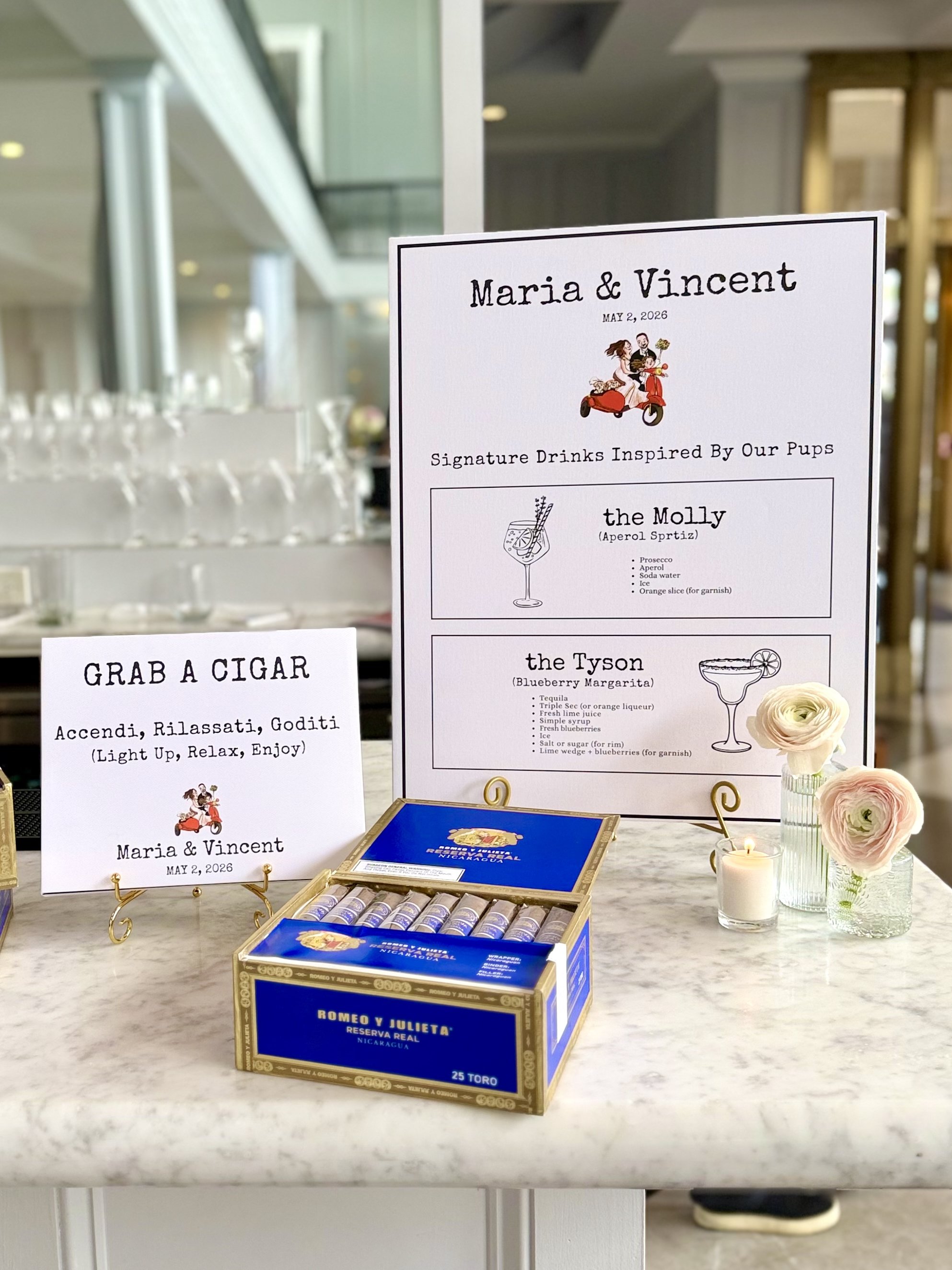 A wedding sign displaying the names Maria and Vincent, with the wedding date May 2, 2026. The sign features a drawing of a couple riding a red scooter. It lists signature drinks inspired by their dogs, including a drink called the Molly with ingredie