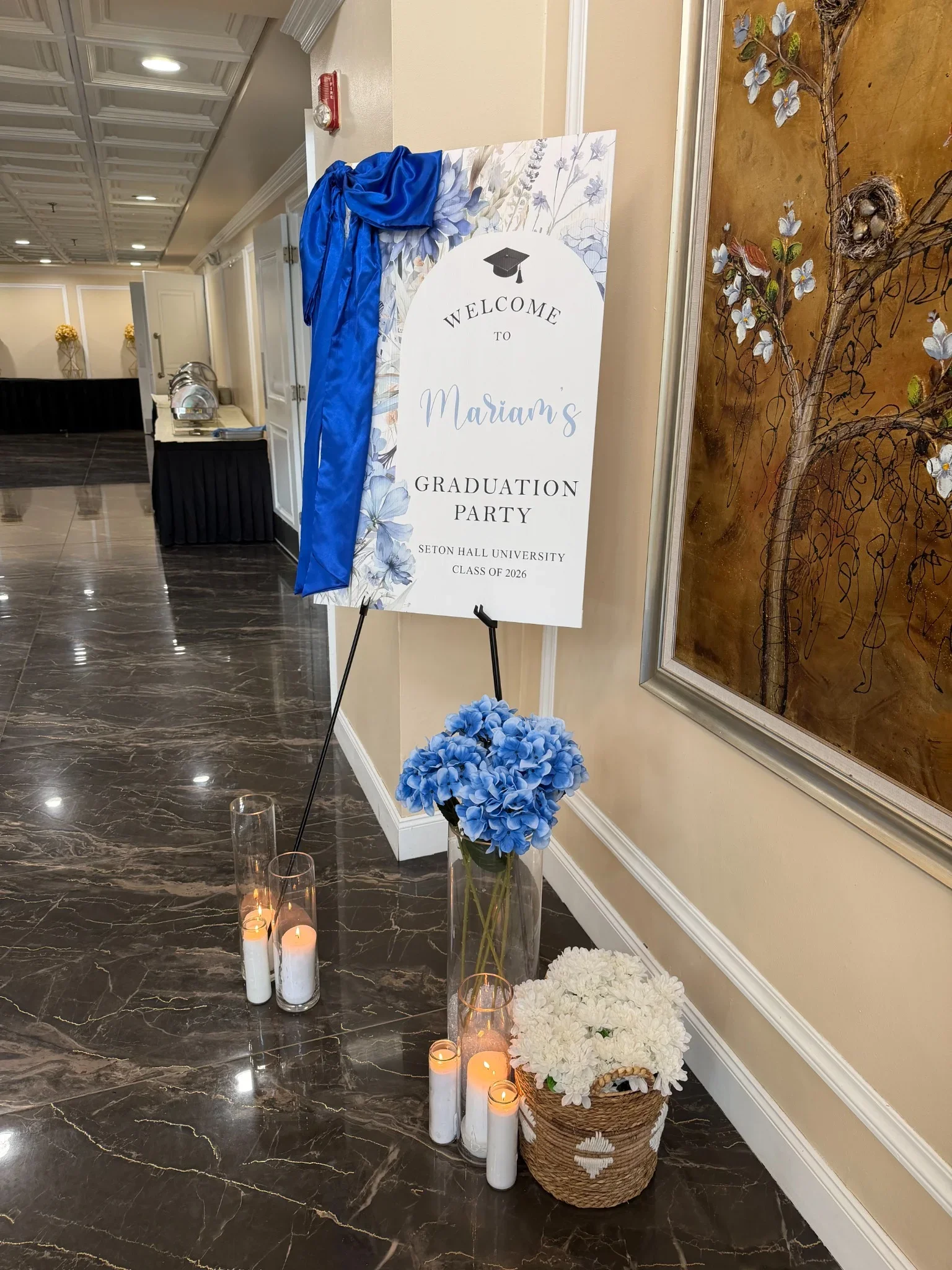 Decorative floral welcome sign for Mariam's graduation party with blue floral accents, blue fabric, candles, and flower arrangements on dark marble floor in an elegant hall.