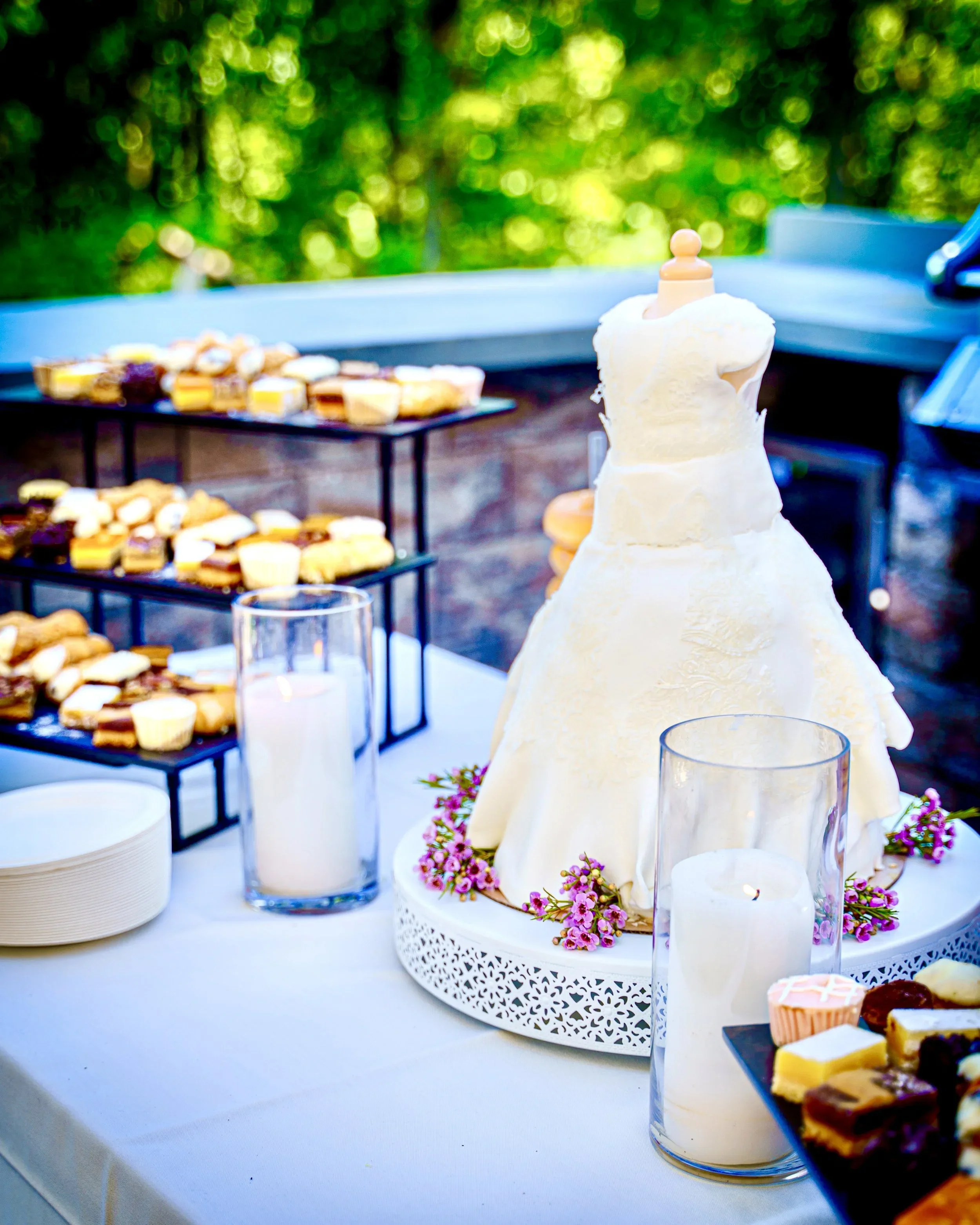 Dessert table with a white cake decorated with pink flowers and surrounded by cupcakes and pastries, outdoors with a vibrant green background.