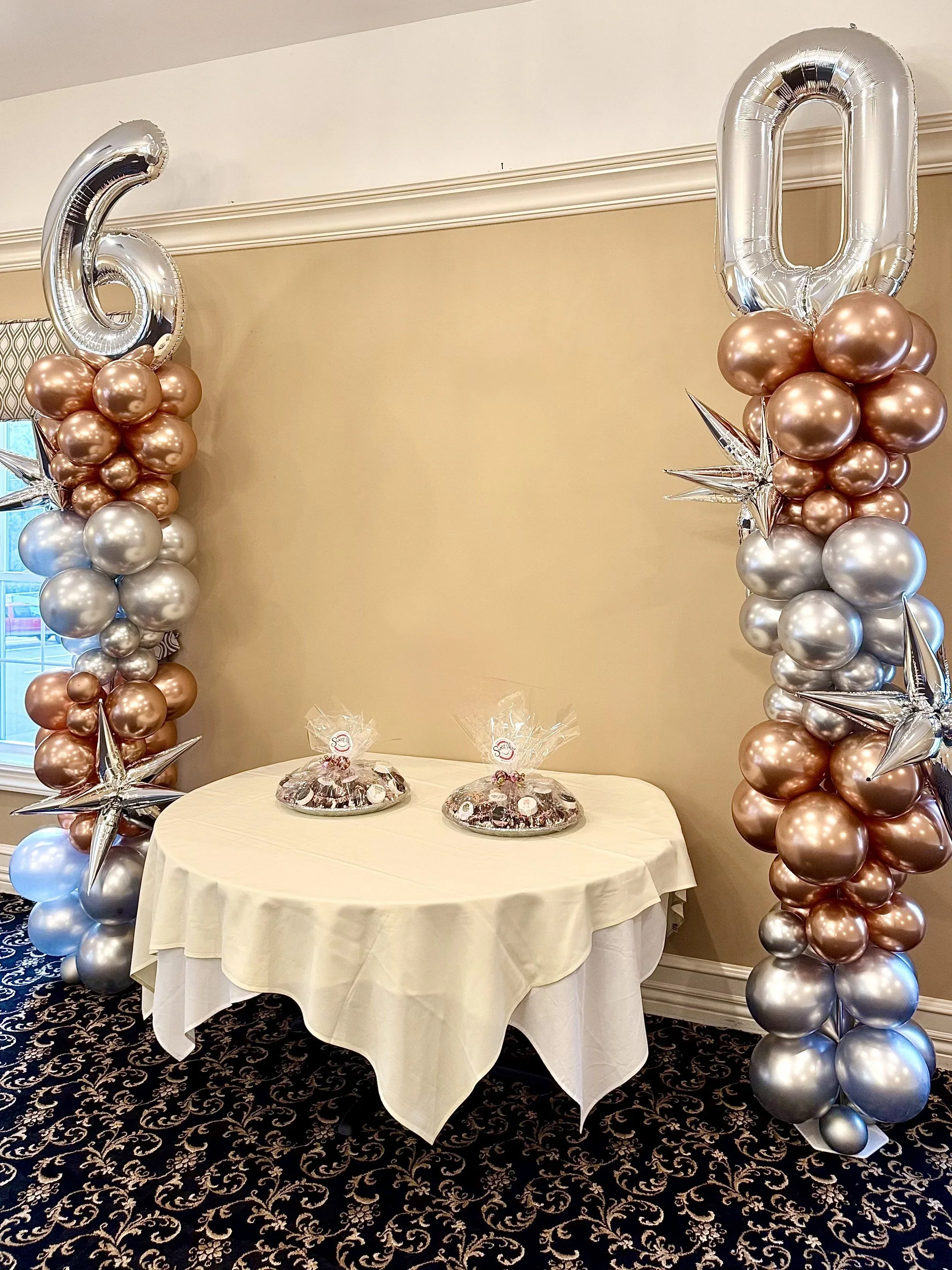 Celebration decorations with 60 and 0 foil balloons, metallic star-shaped balloons, and a table with treats, set in a decorated room with a patterned rug and beige walls.