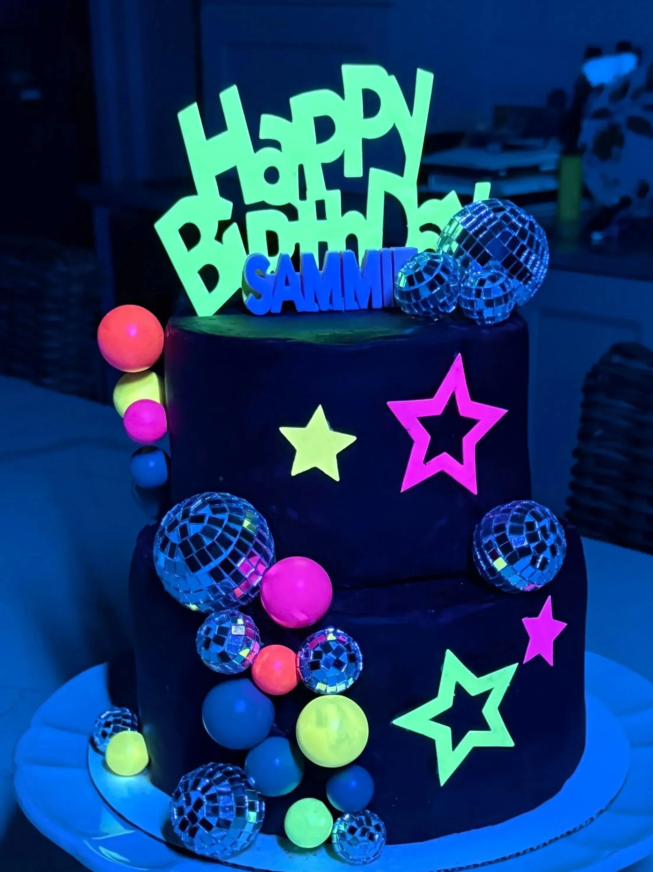 Decorated cake with glowing star and happy birthday sign, adorned with neon-colored and reflective spherical decorations and star cutouts.