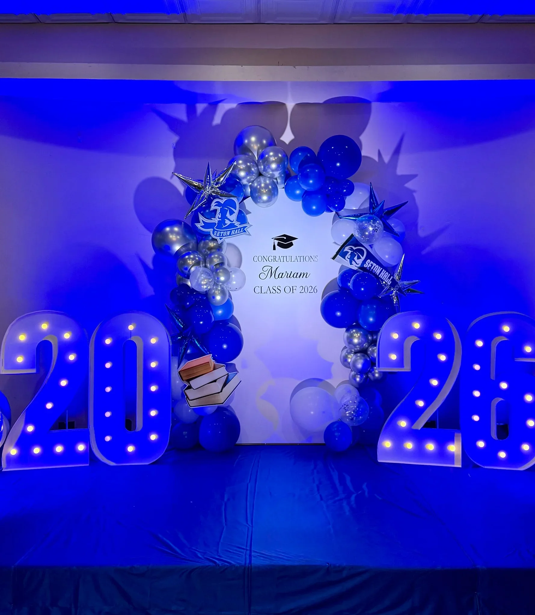 Blue and silver balloons arranged in an arch with star-shaped balloons, graduation caps, and a sign that says "CONGRATULATIONS Maram CLASS OF 2026" with large illuminated numerals "2026" behind them.