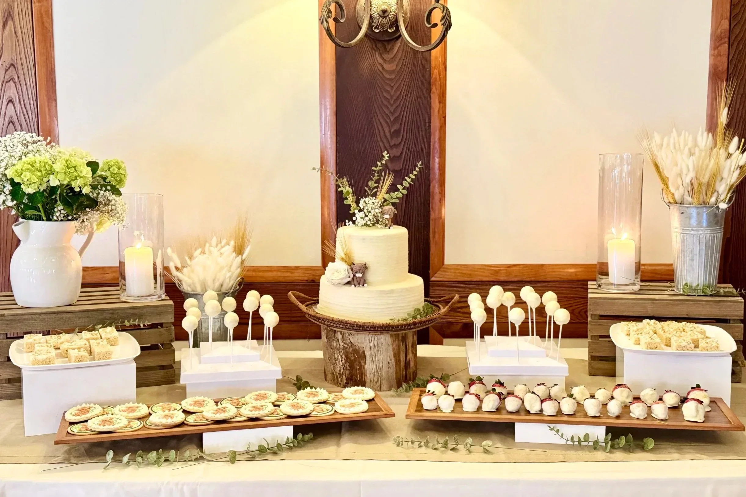 A dessert table with a wedding cake, cookies, cake pops, and cupcakes, decorated with flowers, candles, and greenery, in a rustic style with wooden accents.