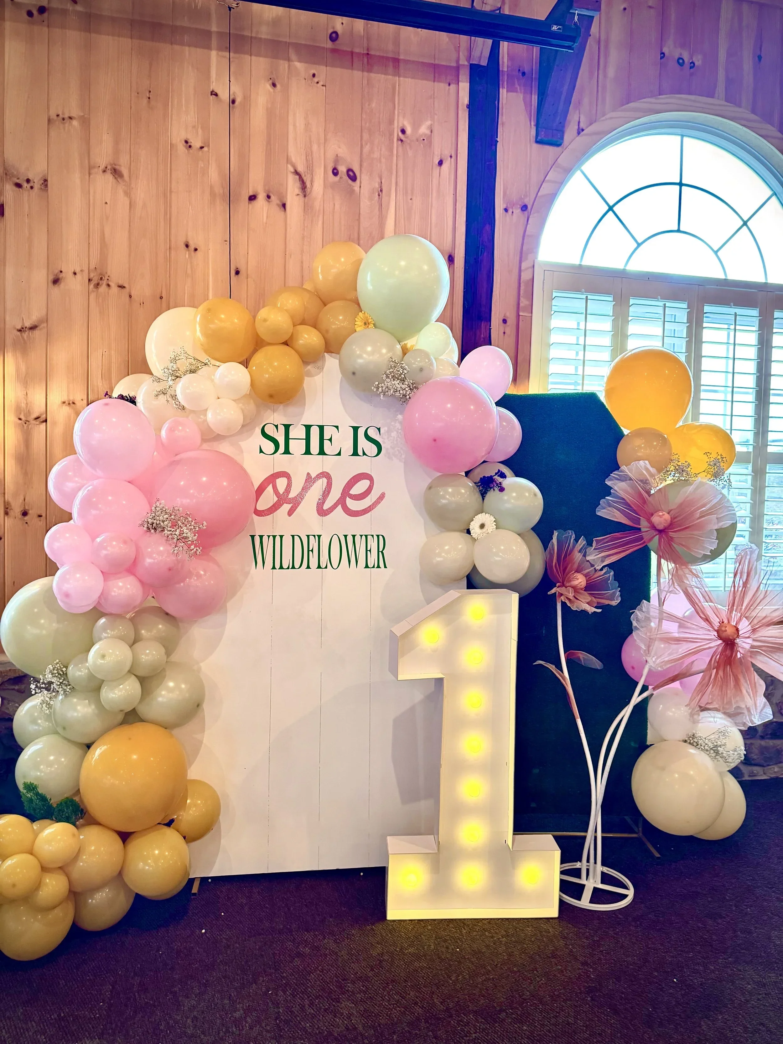 Decorative balloon display for a celebration or event, featuring pastel pink, yellow, and cream balloons, with some floral accents, a sign that reads 'She is one wildflower,' and a lighted letter 'L' on the floor. The background includes a wooden wal