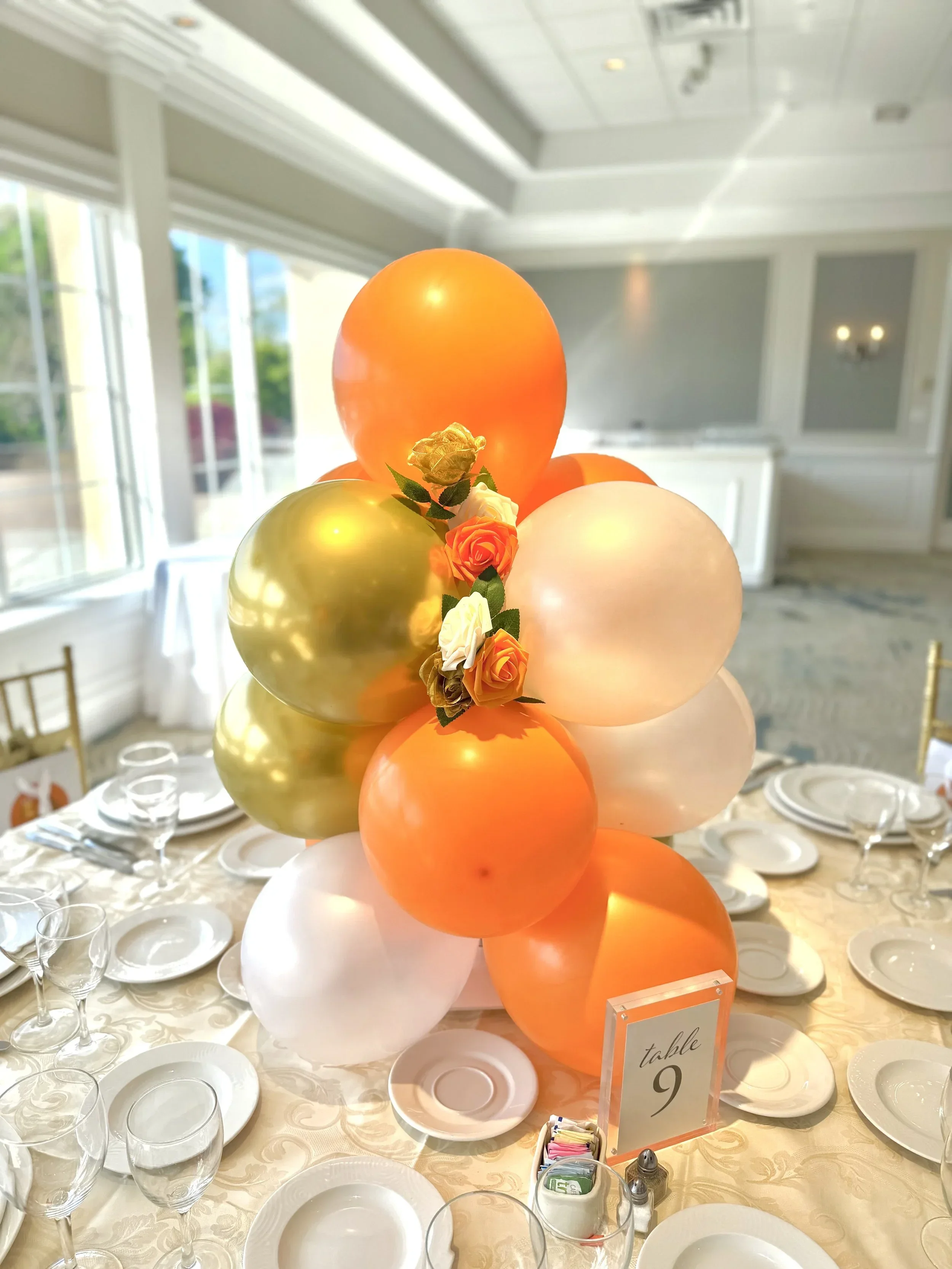 A tall centerpiece made of orange, gold, and pastel-colored balloons with small flowers on a table set for a celebration. The table has white plates, wine glasses, and a table number sign that reads 'table 9'.