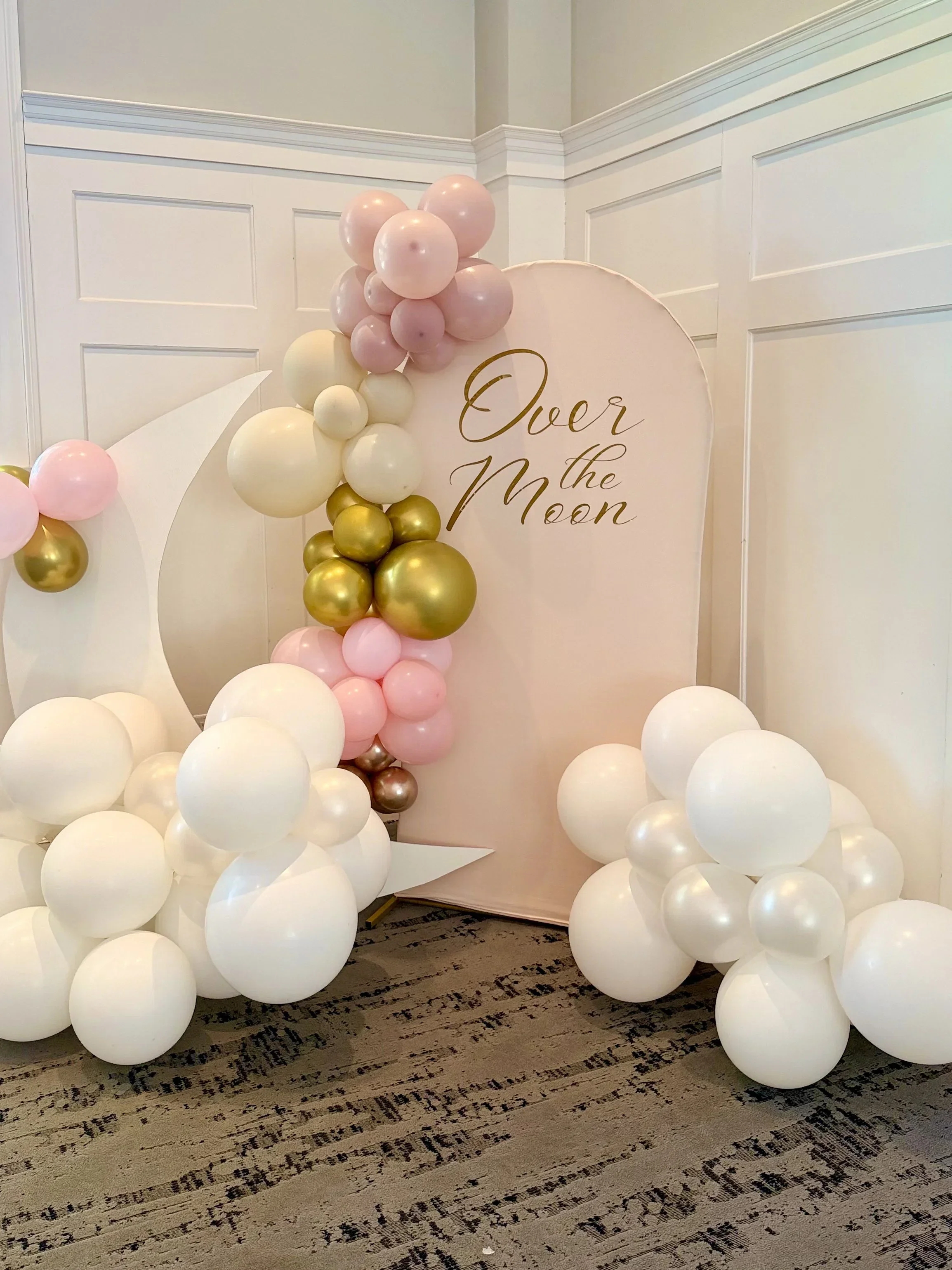 Balloon decoration with pink, white, gold, and beige balloons arranged on a white backdrop with the text "Over the Moon" in gold lettering.