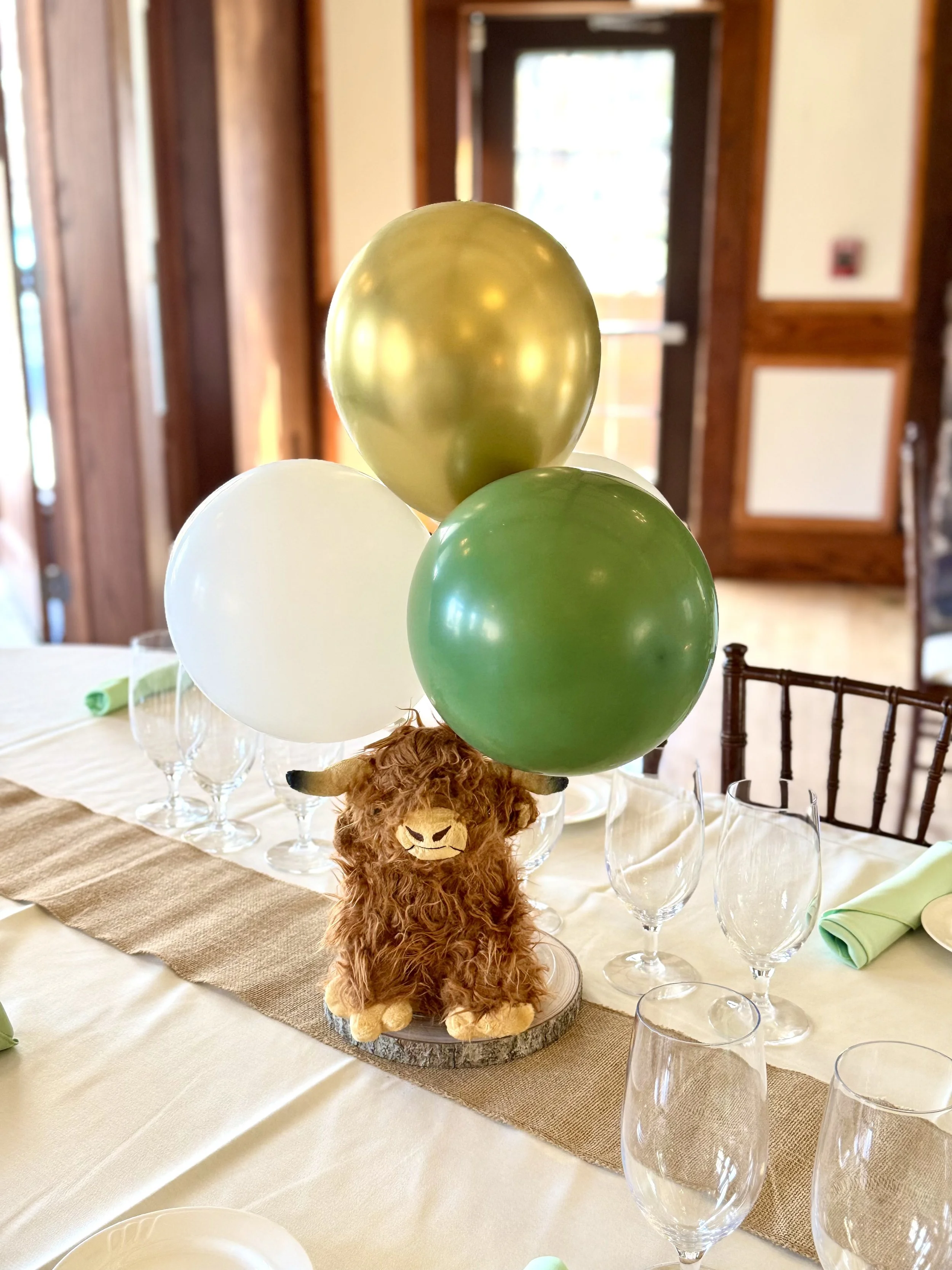 Decorative centerpiece with plush llama and green, white, and gold balloons on a banquet table set for a celebration or party.
