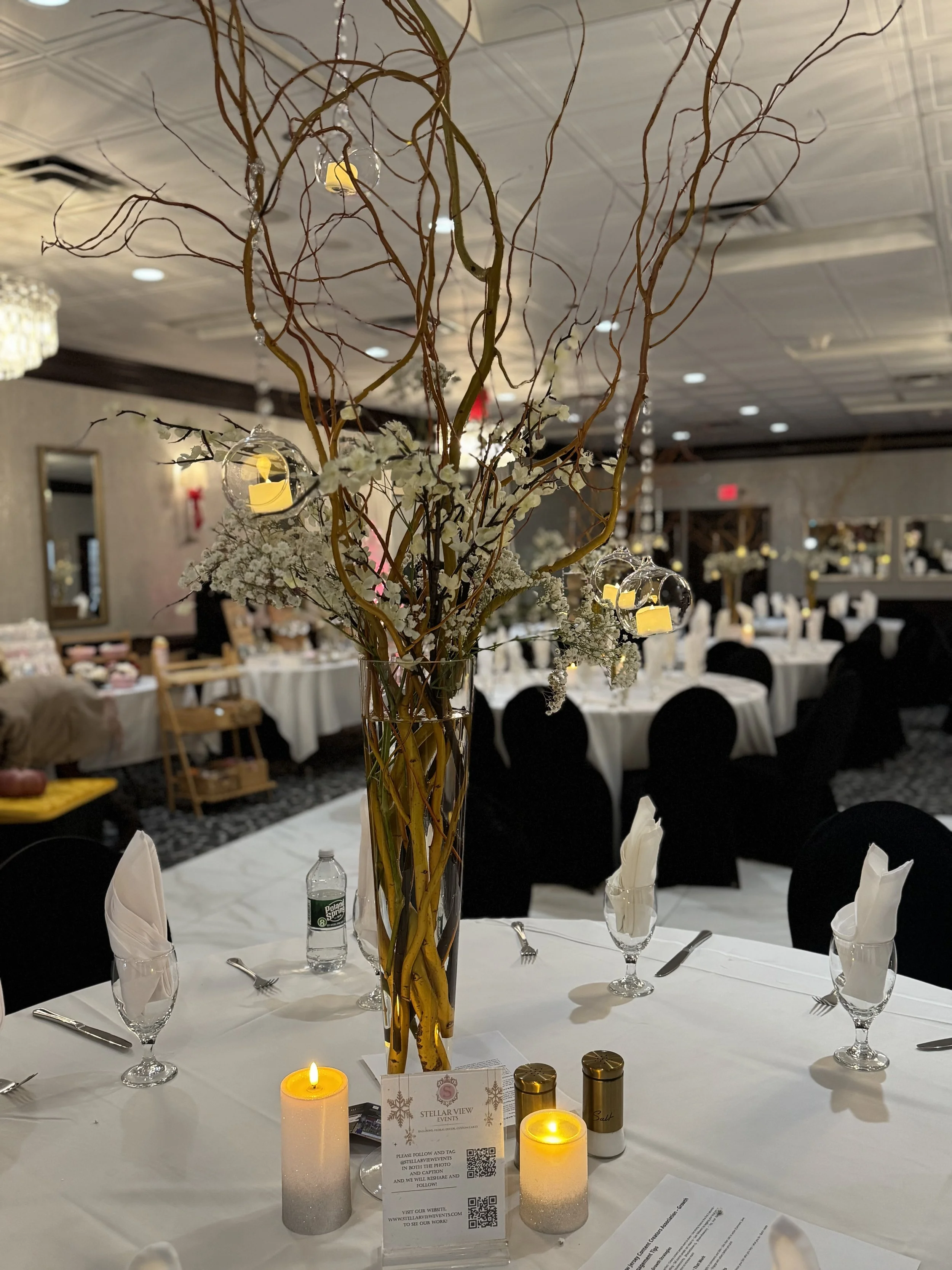 A banquet table decorated with a tall centerpiece of twisted branches, white flowers, hanging ornaments, and small candles. The table is set with white napkins, silverware, wine glasses, and a bottle of water.