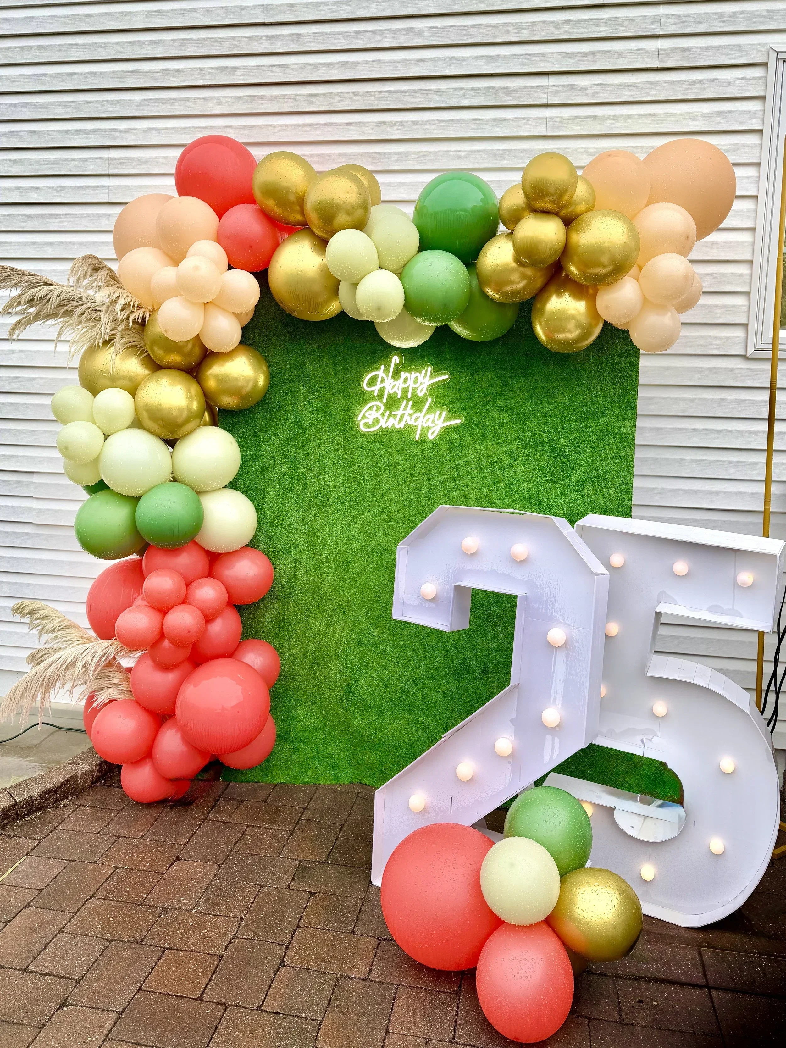 Colorful birthday decoration featuring balloons in shades of red, gold, green, peach, and cream, arranged around a green backdrop with a neon 'Happy Birthday' sign and a large illuminated white number '2' with light bulbs, set on a brick pavement.