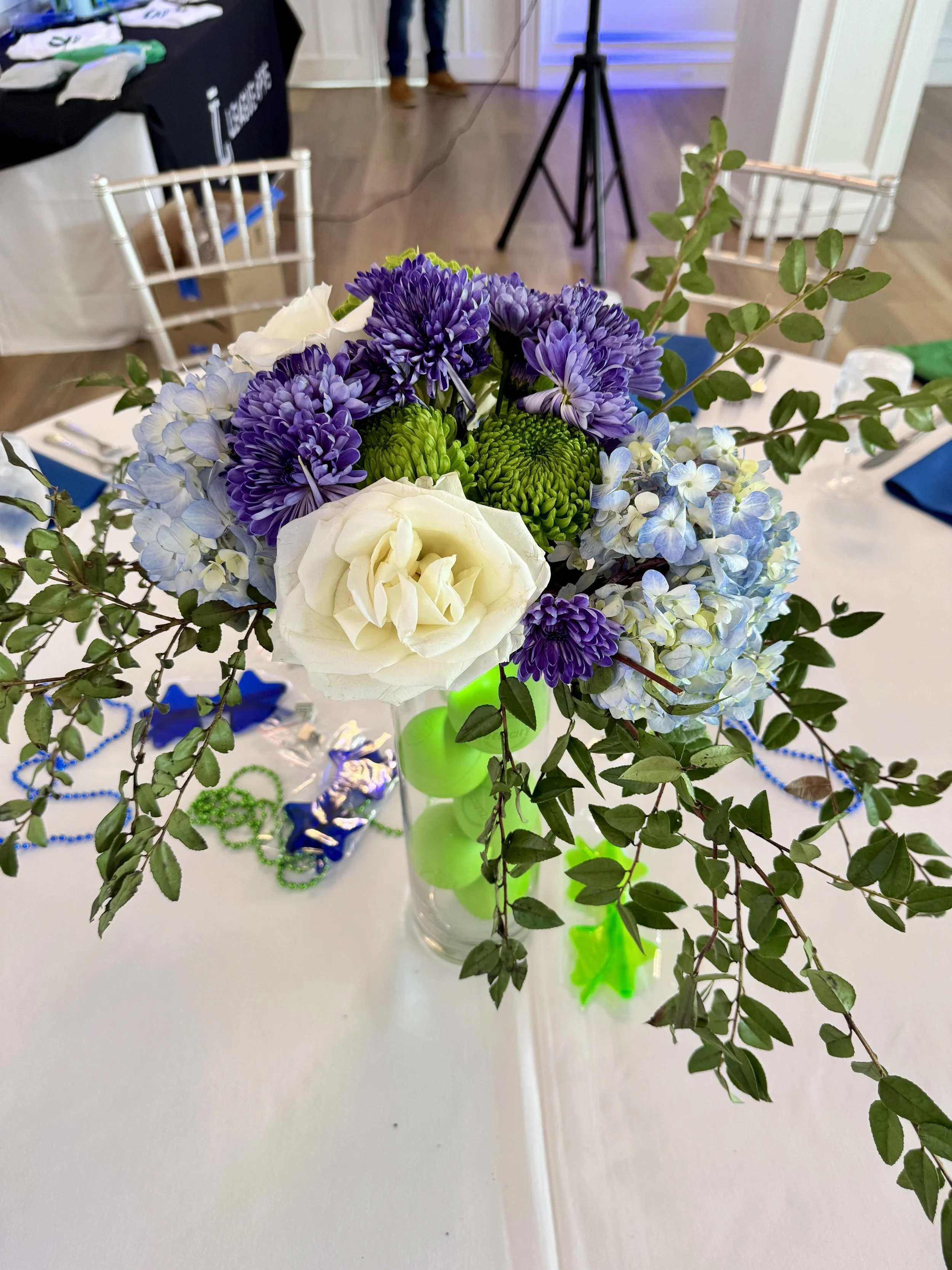 Close-up of a floral centerpiece with white roses, purple chrysanthemums, light blue hydrangeas, and green accents on a table at an event.