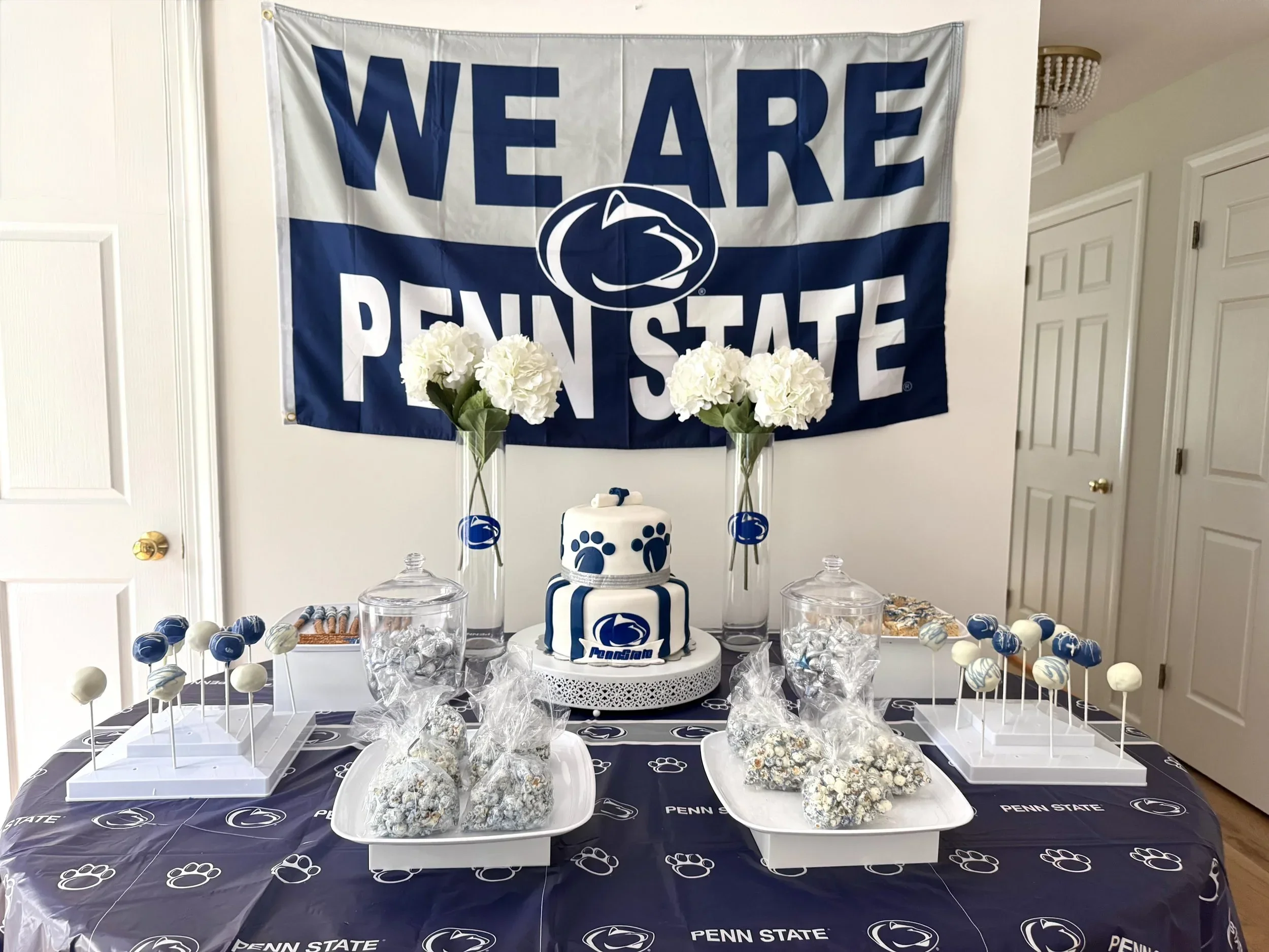 Decorative table setup for Penn State with a Penn State themed cake, white and blue cake pops, floral arrangements, and penn state banners.