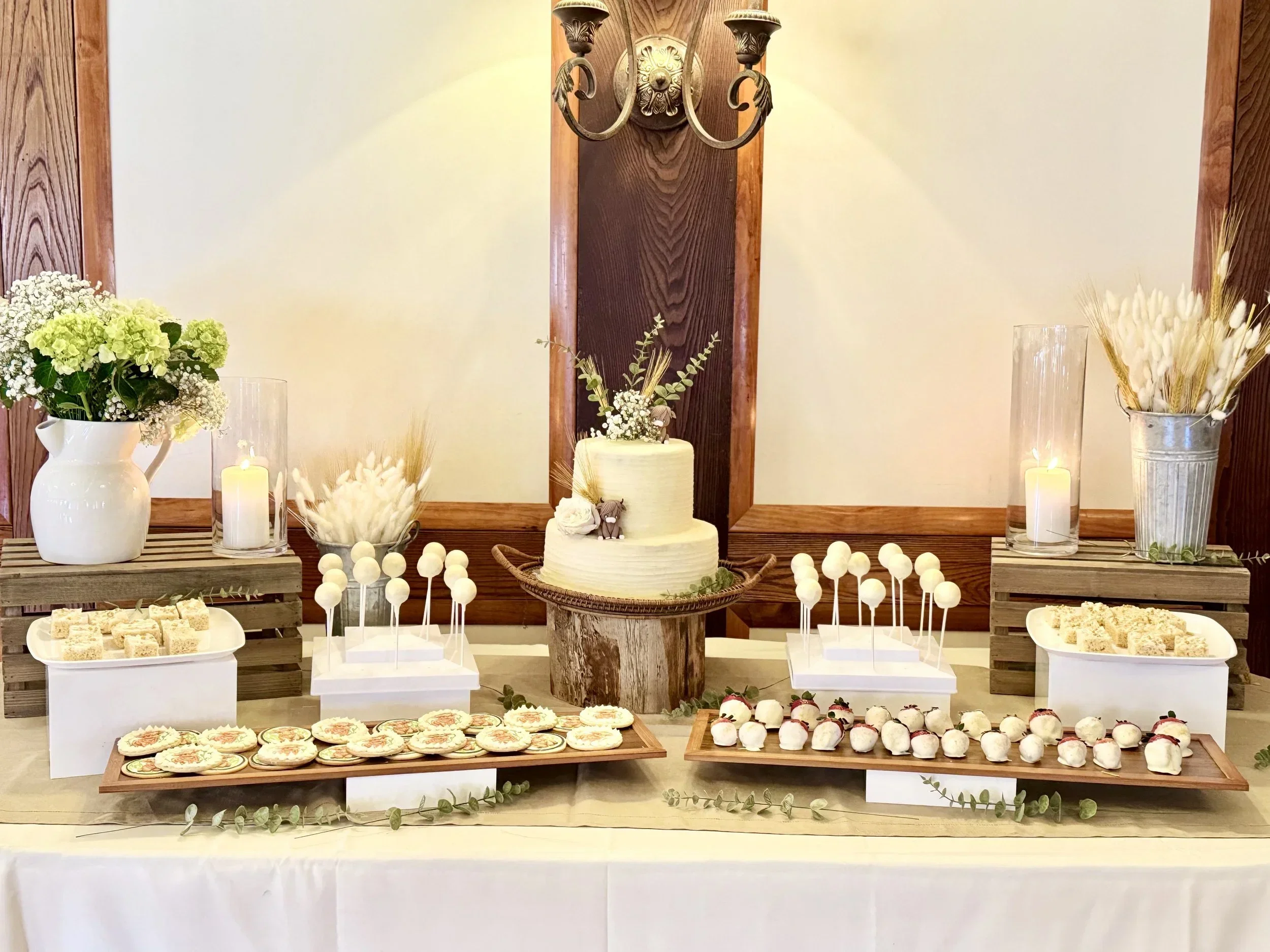 Wedding dessert table with a two-tier white cake, assorted cookies, strawberries, and decorated with faux animals, surrounded by flowers and candles, against a wooden wall with a chandelier.