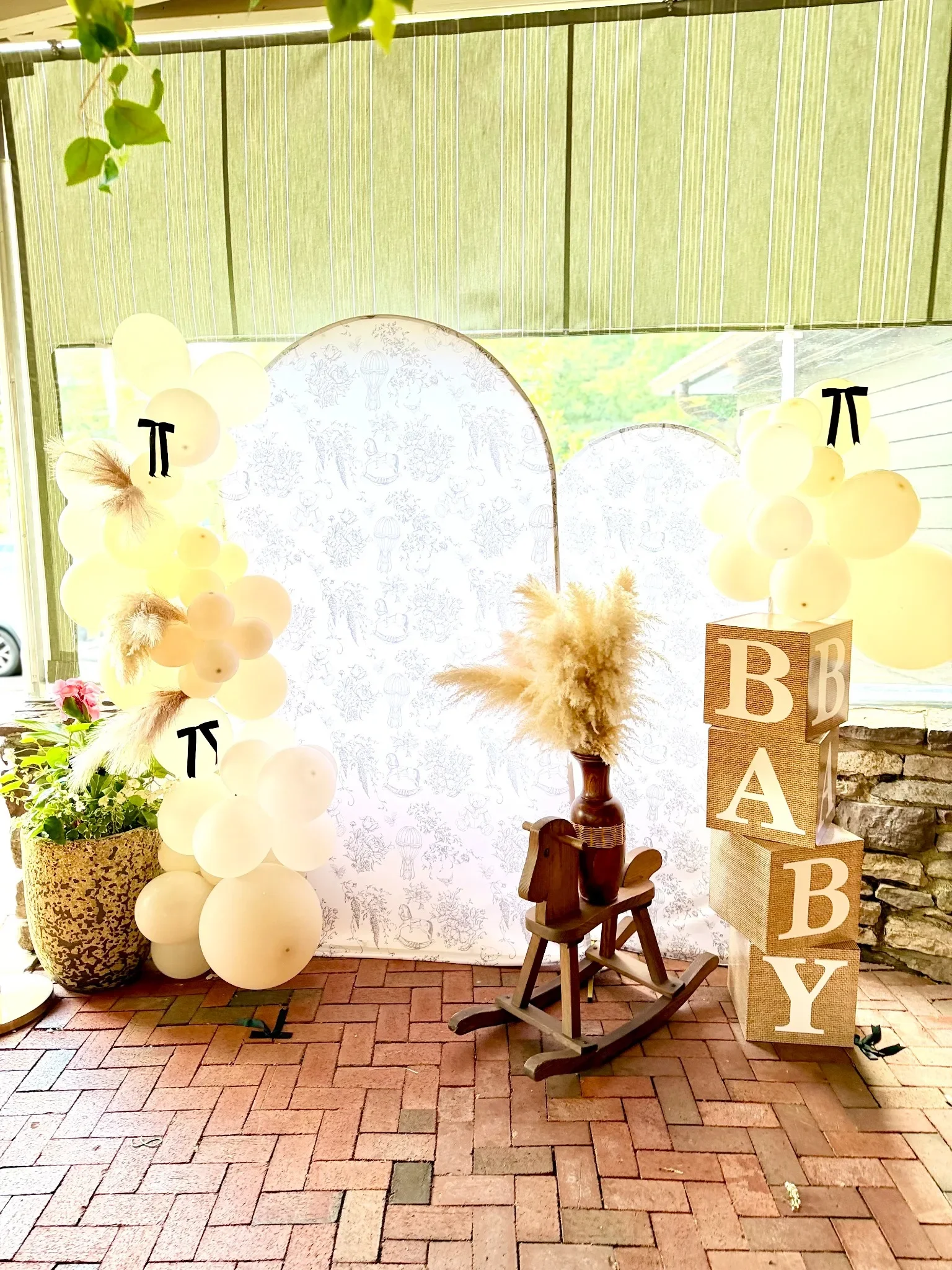 Decorative display with cream-colored balloons, a potted plant, a wooden rocking horse, and wooden blocks spelling 'BABY' on a brick patio with a sheer curtain backdrop.