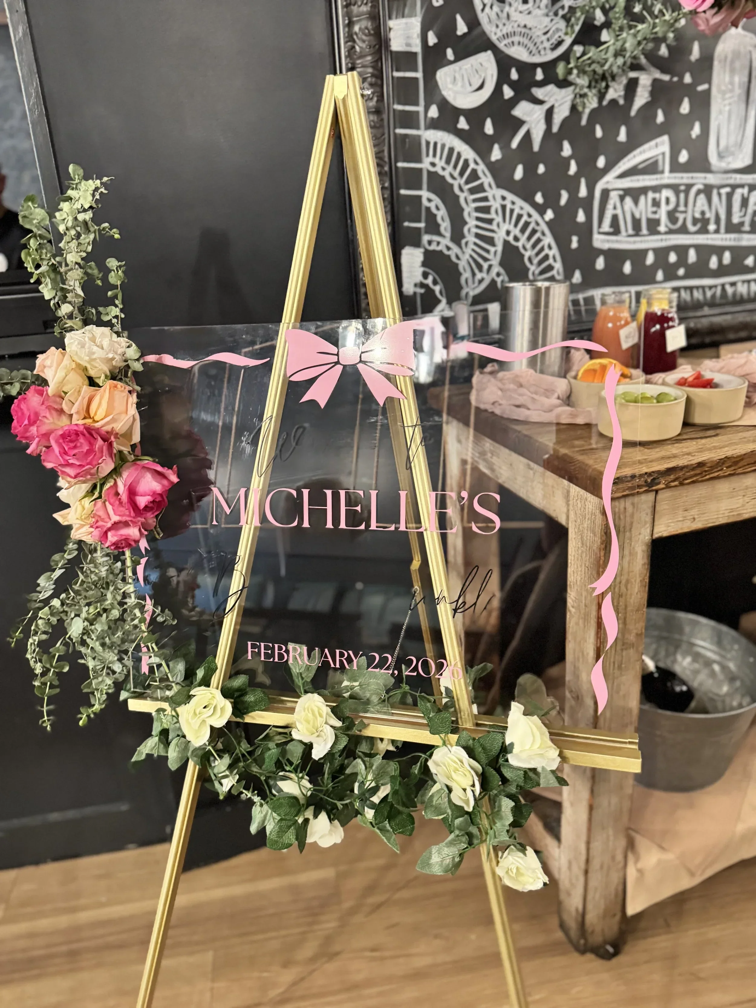 A floral display with a gold easel holding a sign that reads 'MICHELLE'S' and a date 'FEBRUARY 22, 2026.' The display is decorated with pink and white flowers and green foliage, and is set against a black chalkboard with white chalk illustrations and