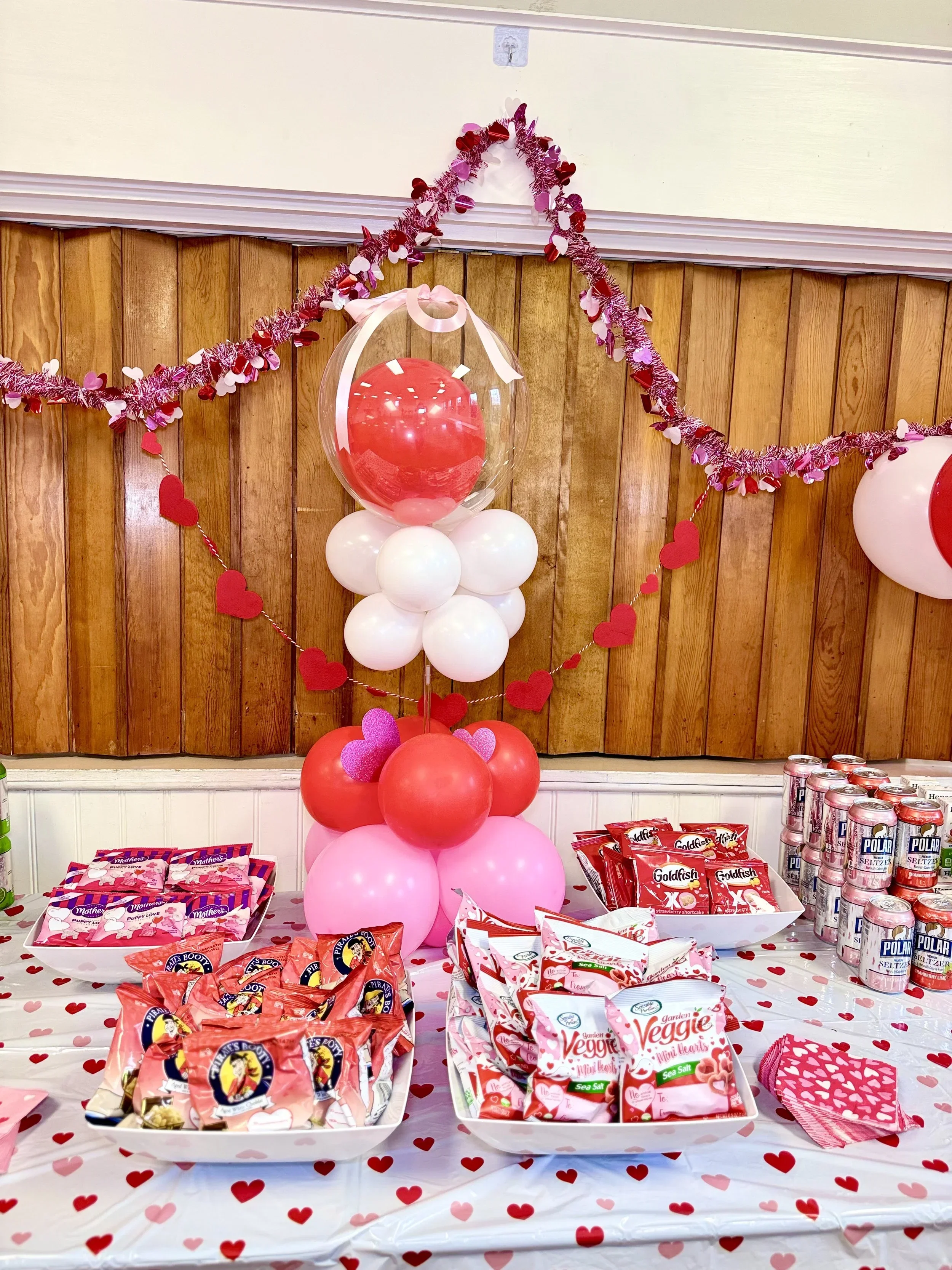 Valentine's Day themed snack table decorated with pink and red balloons, heart-shaped garland, and various snack packages including chips and candy, set against a wooden-paneled wall.