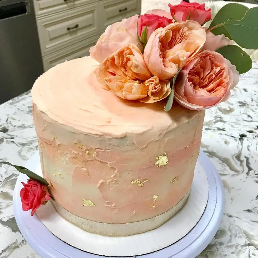 A round pastel pink cake with gold leaf decor, topped with a bouquet of peach and pink peonies, placed on a marble surface.