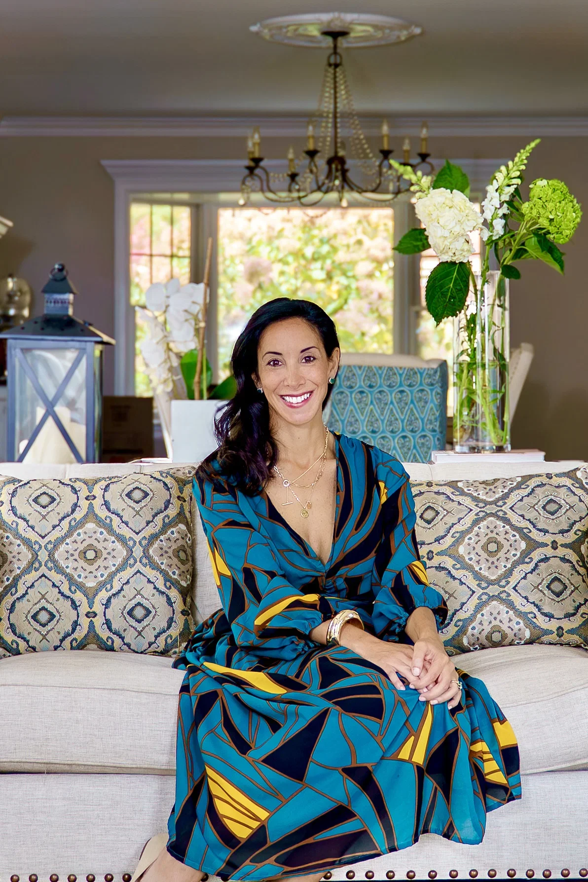 A woman sitting on a patterned sofa in a well-lit living room with a large window, green plants, and decorative items in the background.