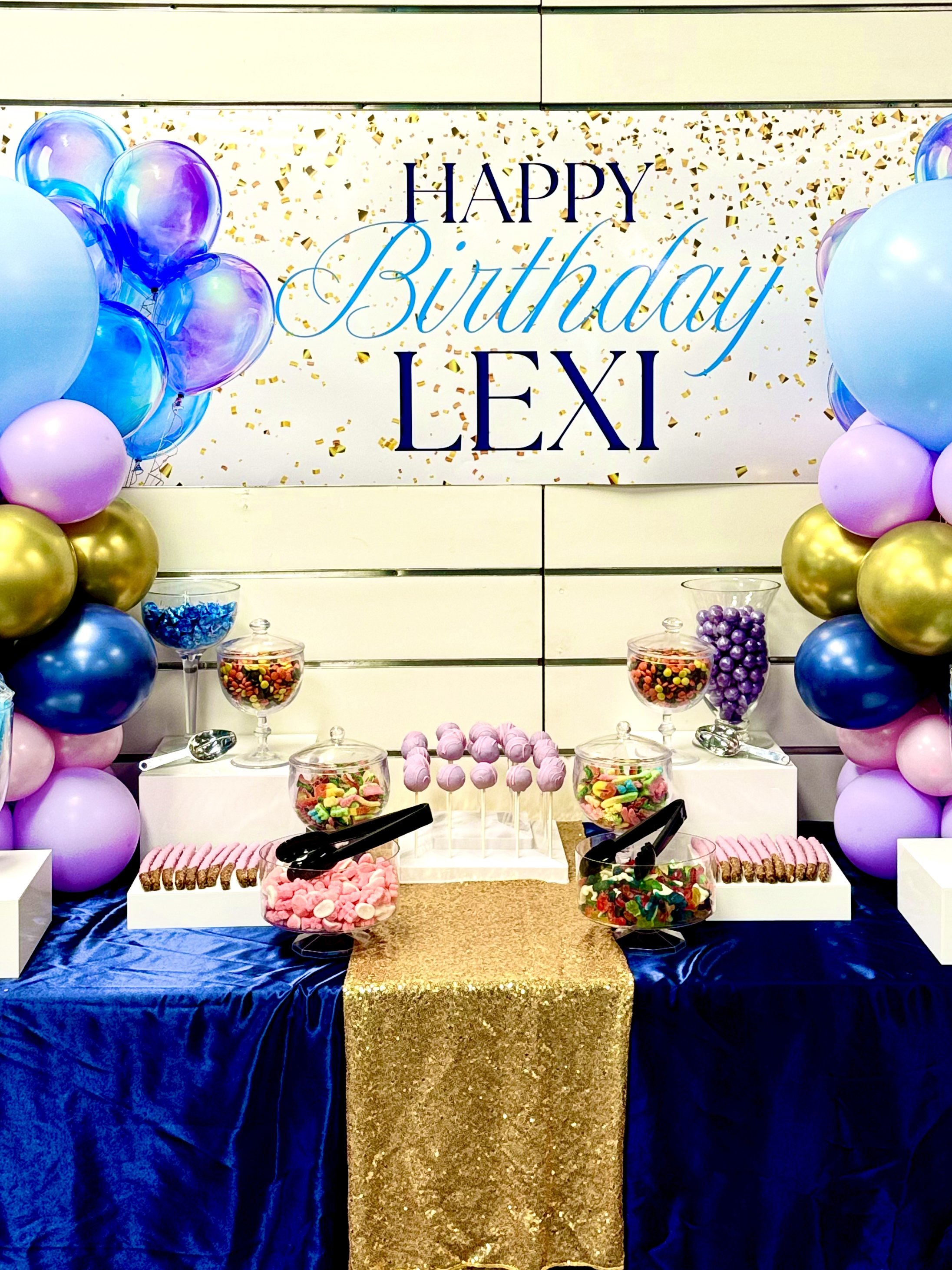 Decorative candy table with balloons, sweets, and a 'Happy Birthday Lexi' sign.