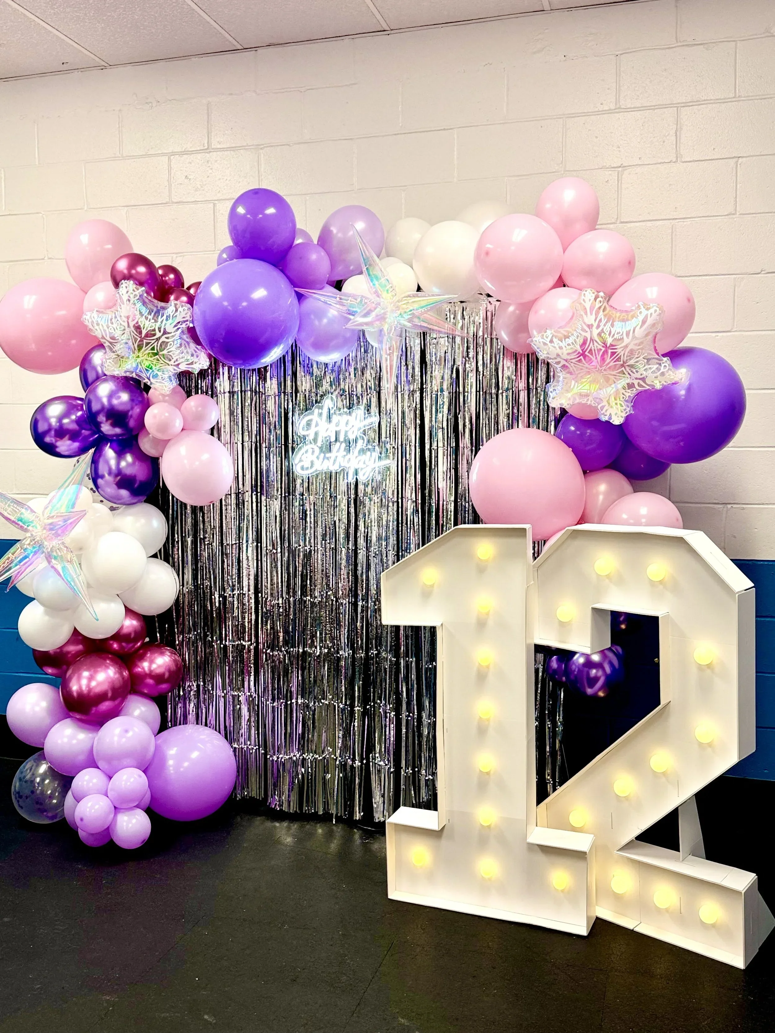 Decorative photo booth area with pink, purple, and white balloons, holographic star balloons, metallic fringe background, illuminated 'A' sign, and a large light-up 'LOVE' sign.
