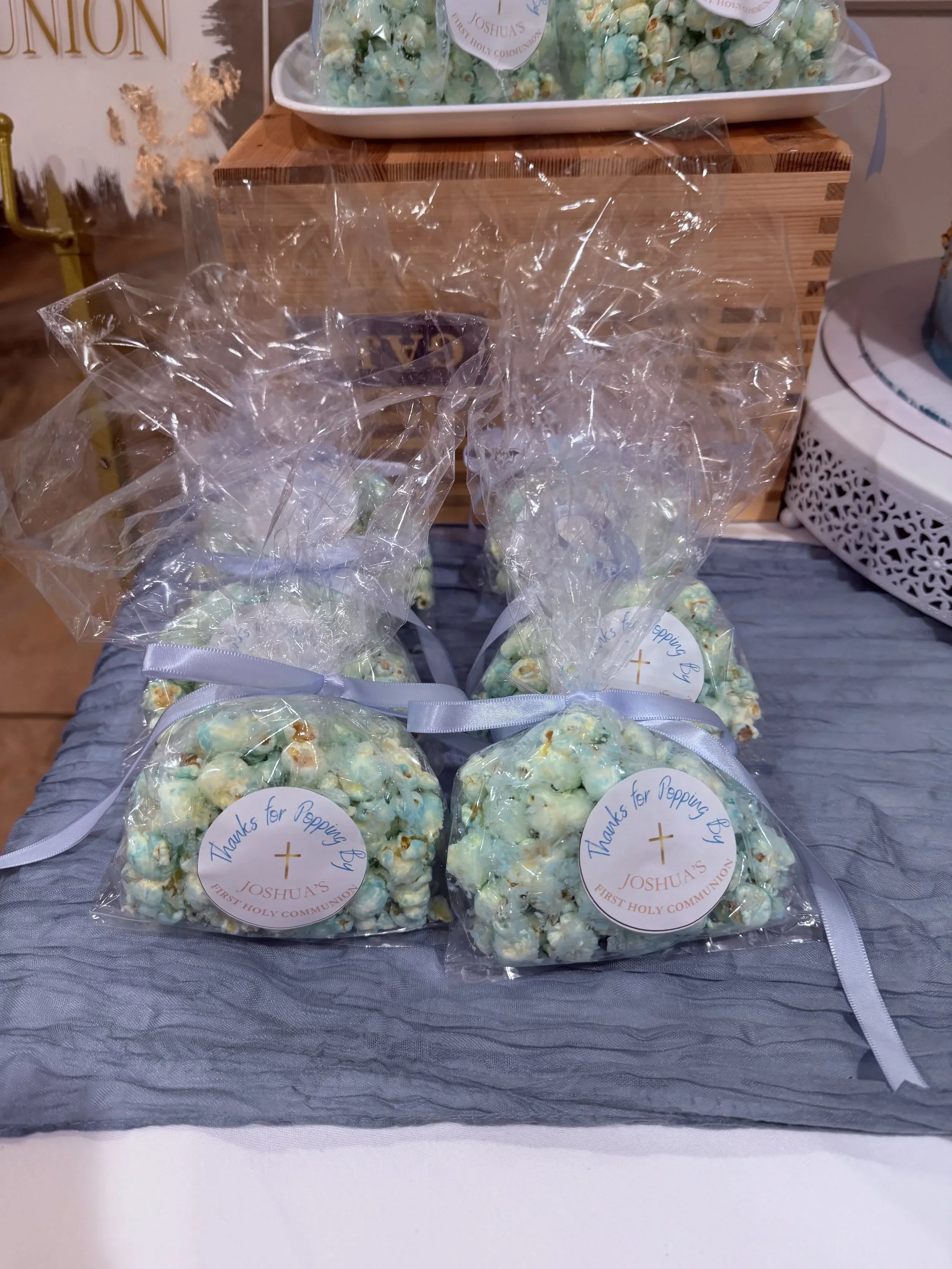 Bags of popcorn with pastel green and white colors, tied with white ribbon, placed on a table and labeled with a round tag that says 'Thanks for Popping By' and 'Joshua's First Holy Communion'.