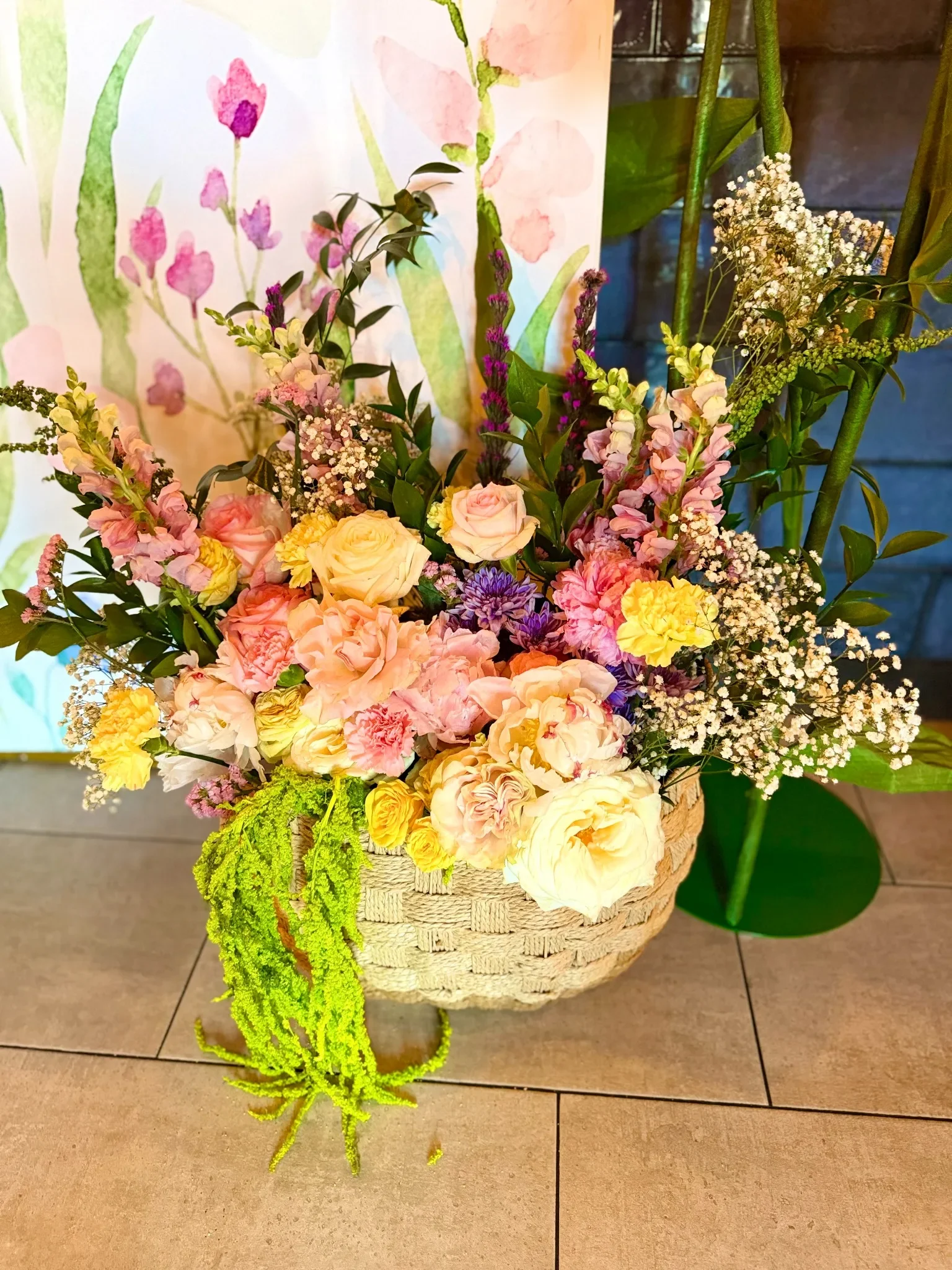 A large basket filled with a colorful arrangement of roses, carnations, astilbe, and baby's breath on a tiled floor.