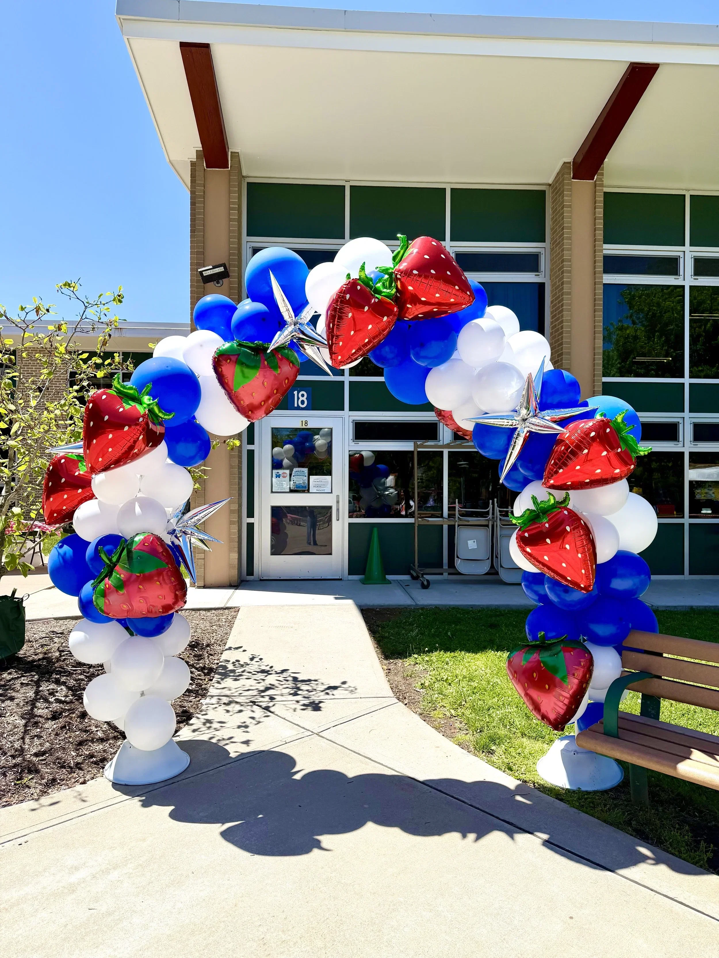 Colorful balloon arch with red strawberries, white, blue, and silver balloons outside a building decorated for a celebration.