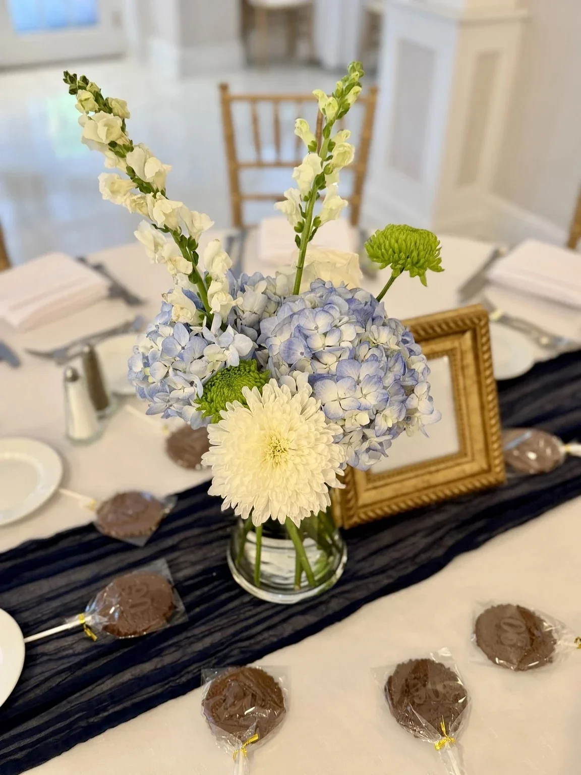 A floral centerpiece with white and light purple flowers in a glass vase on a white table, decorated with a black cloth runner, small chocolates wrapped in clear plastic, a gold picture frame, and place settings in a decorated room.