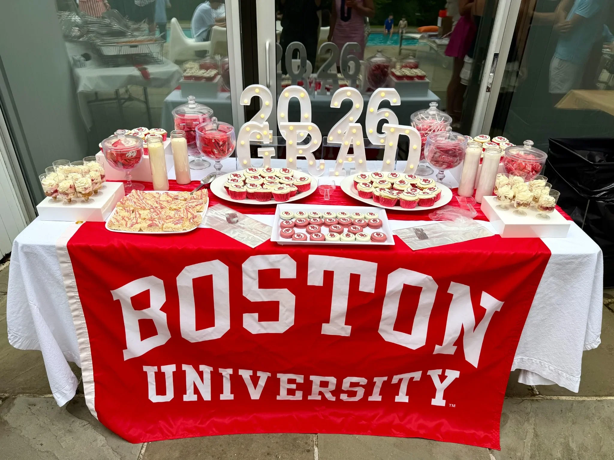 A table with red and white decorations displaying desserts including cupcakes with a university logo, and jars of candy. Behind the table, there are people near a swimming pool and the table has a large red cloth with the words 'University' printed o