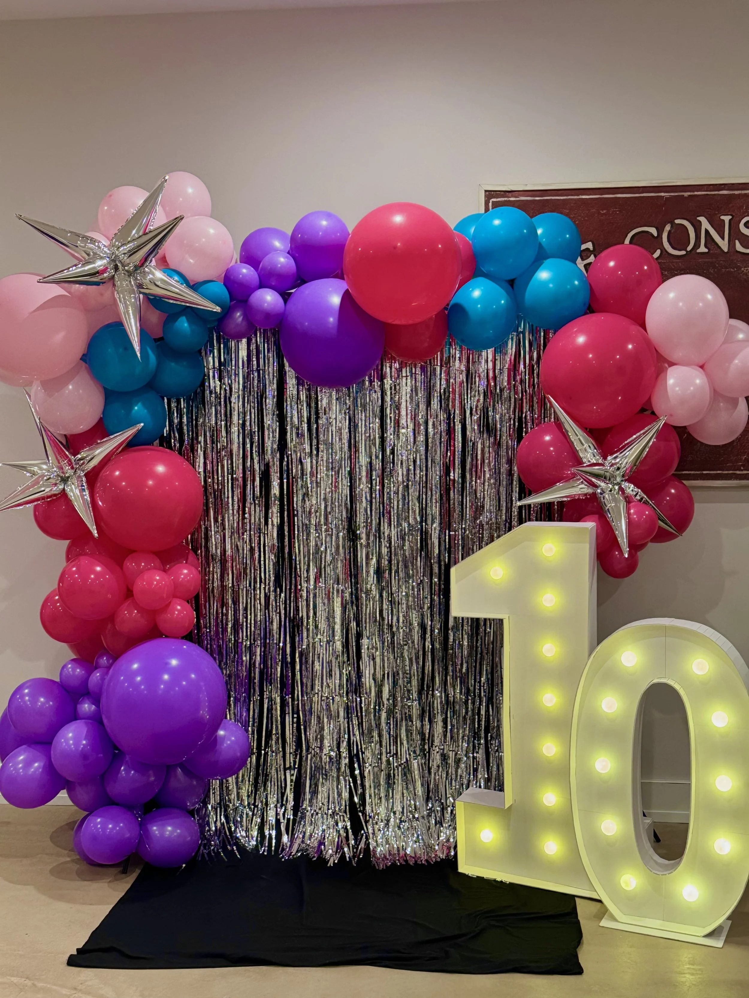 Colorful balloon arch with pink, purple, blue, and red balloons, silver star-shaped balloons, silver tinsel backdrop, and a large illuminated number 10 for celebration.