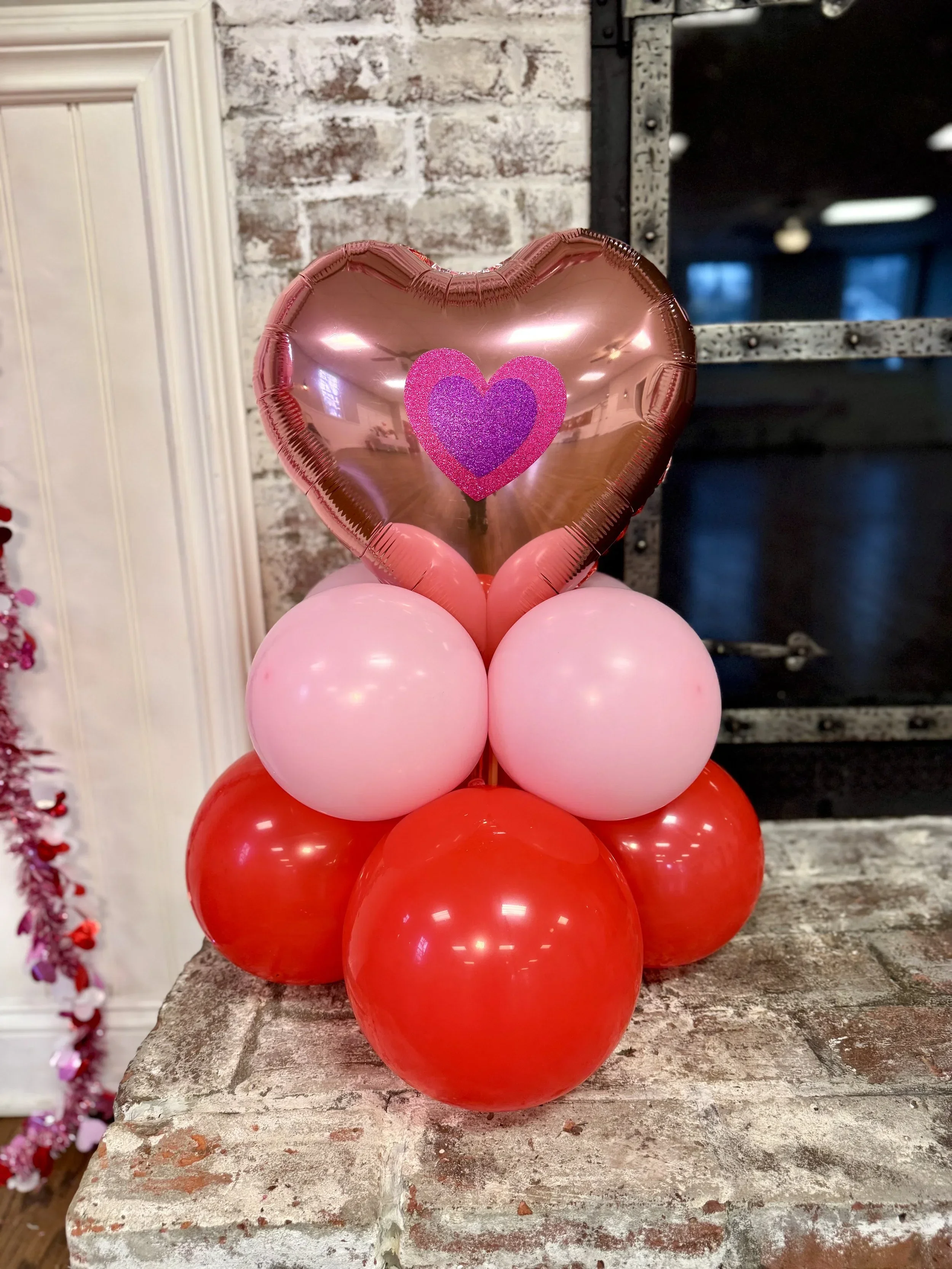 A cluster of pink and red balloons with a pink heart-shaped balloon on top, displayed on a rustic wooden surface with a brick wall background.
