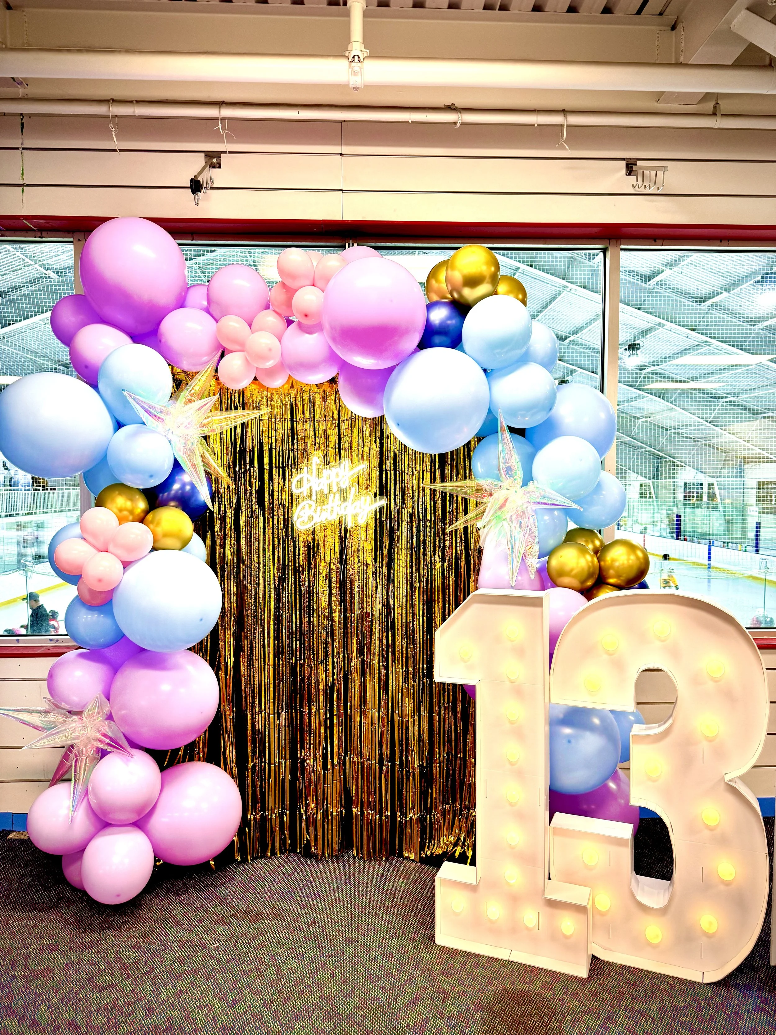 Colorful birthday decoration featuring balloons in pink, purple, blue, gold, and star-shaped balloons, with a large illuminated number 13 and a golden and black metallic curtain backdrop inside an ice skating rink.