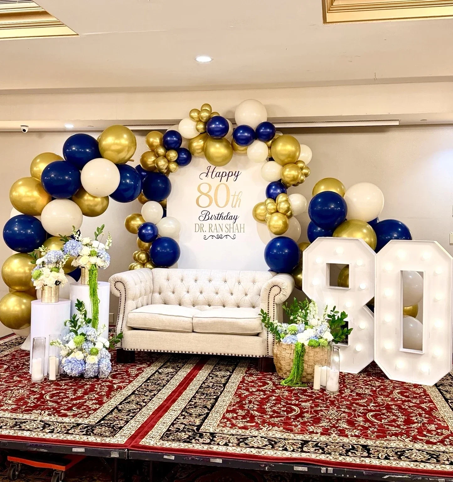Decorative stage with a white tufted sofa, floral arrangements, a backdrop with balloons in gold, navy blue, and white, and a sign celebrating an 80th birthday for Dr. Ranshah.
