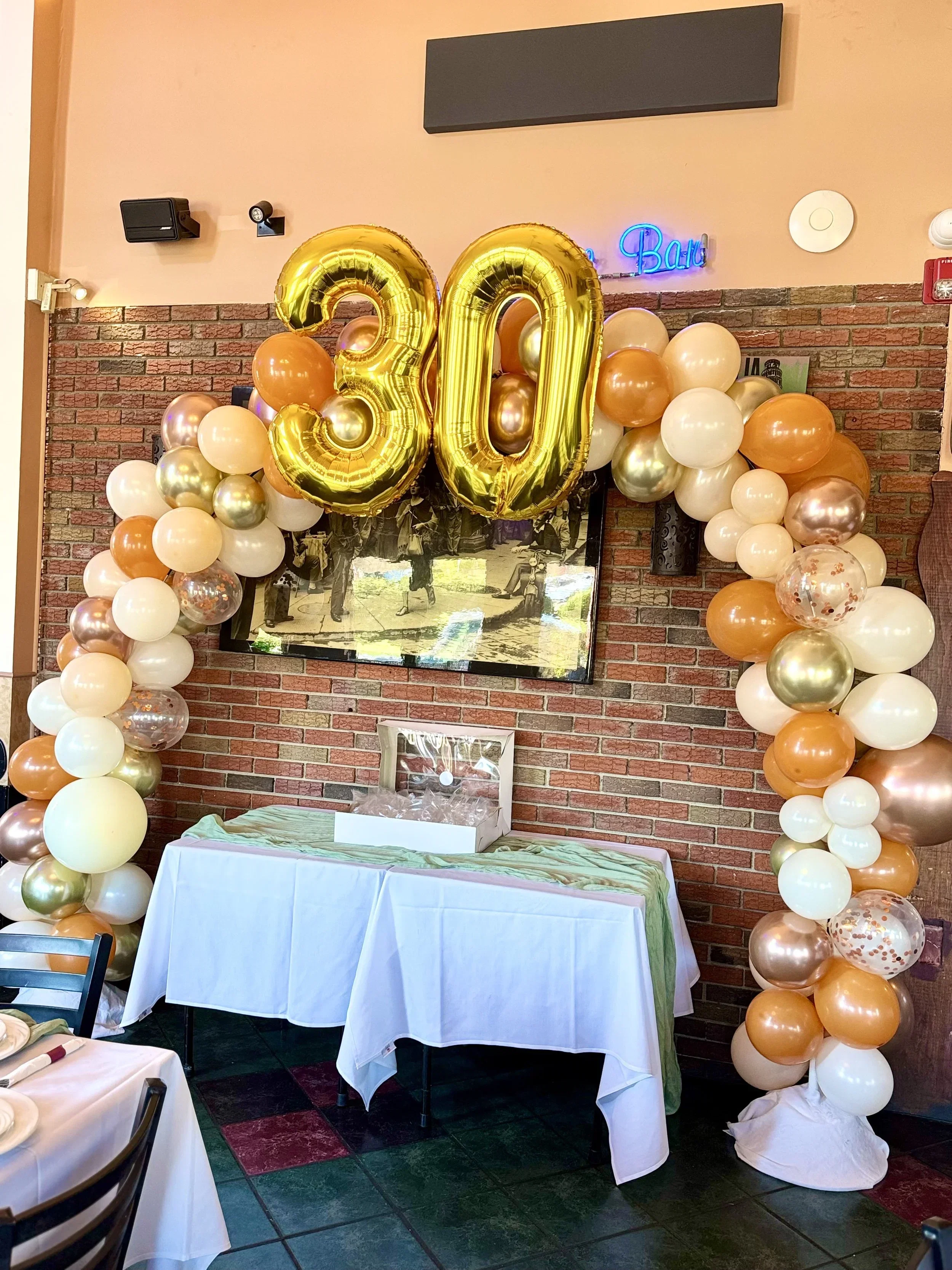 Decorations for a 20th birthday celebration including large gold balloons forming the number 20, surrounded by gold, white, and orange balloons arranged in an arch on a brick wall, with a table draped in white cloth and a green runner underneath.